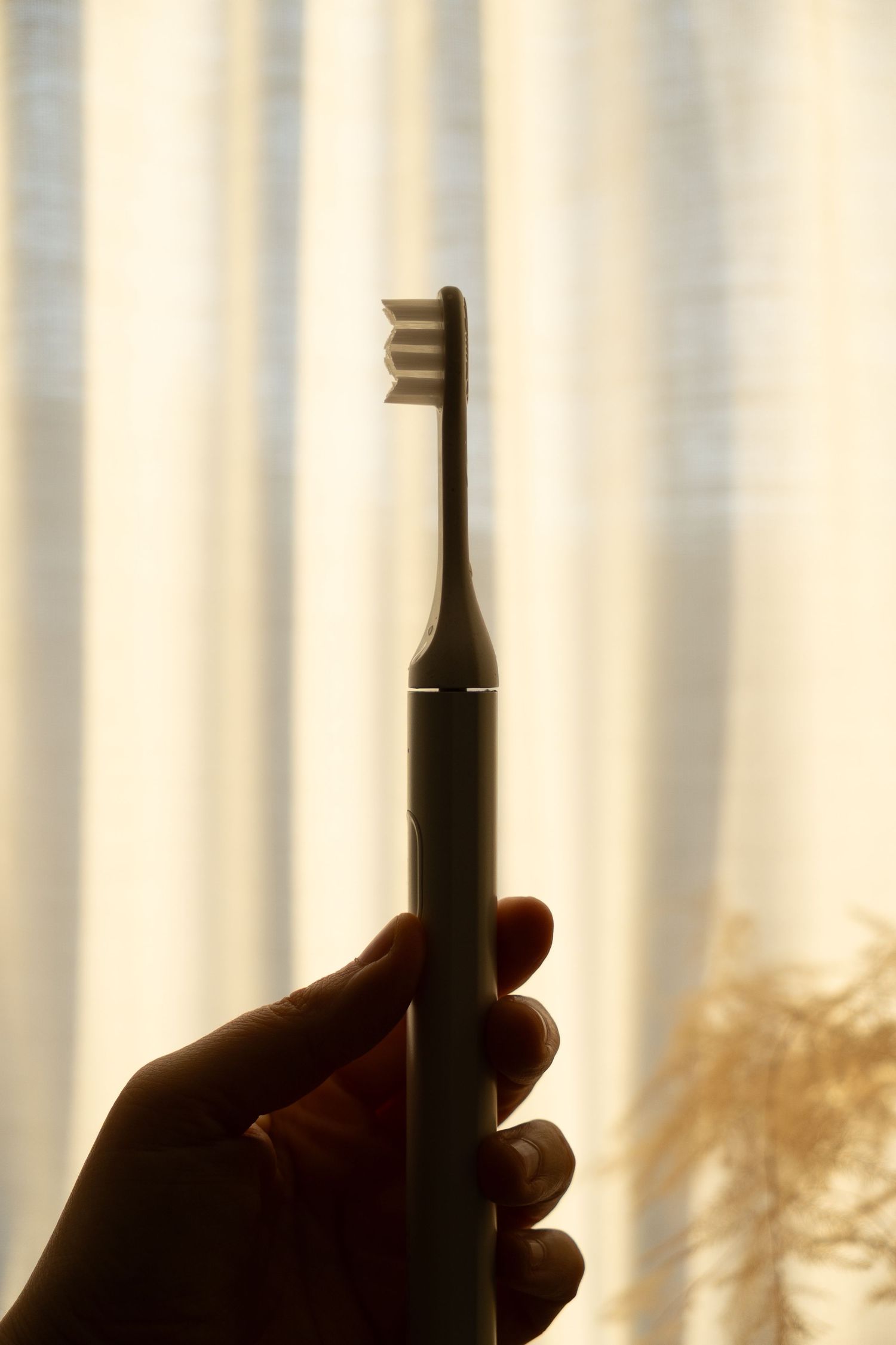 Silhouette of a hand holding an electric toothbrush against a warm backlit curtain.