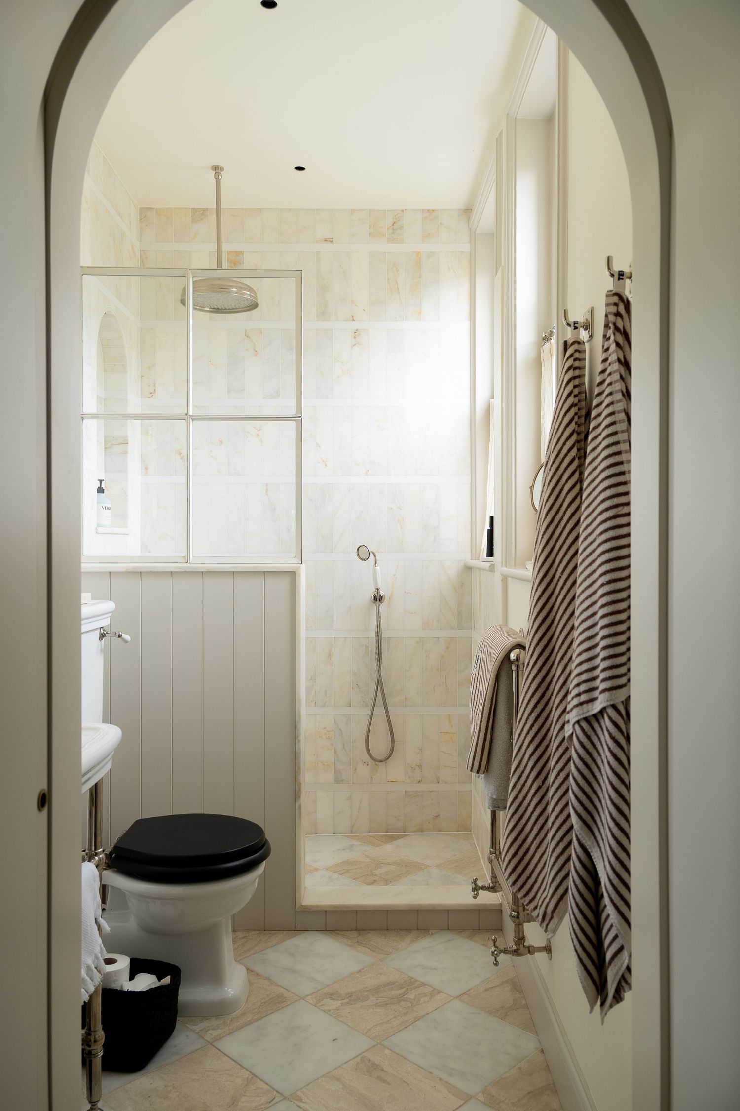 Elegant white bathroom with arched doorway, black toilet seat, walk-in shower, marble tiles, and striped towels on heated rail.