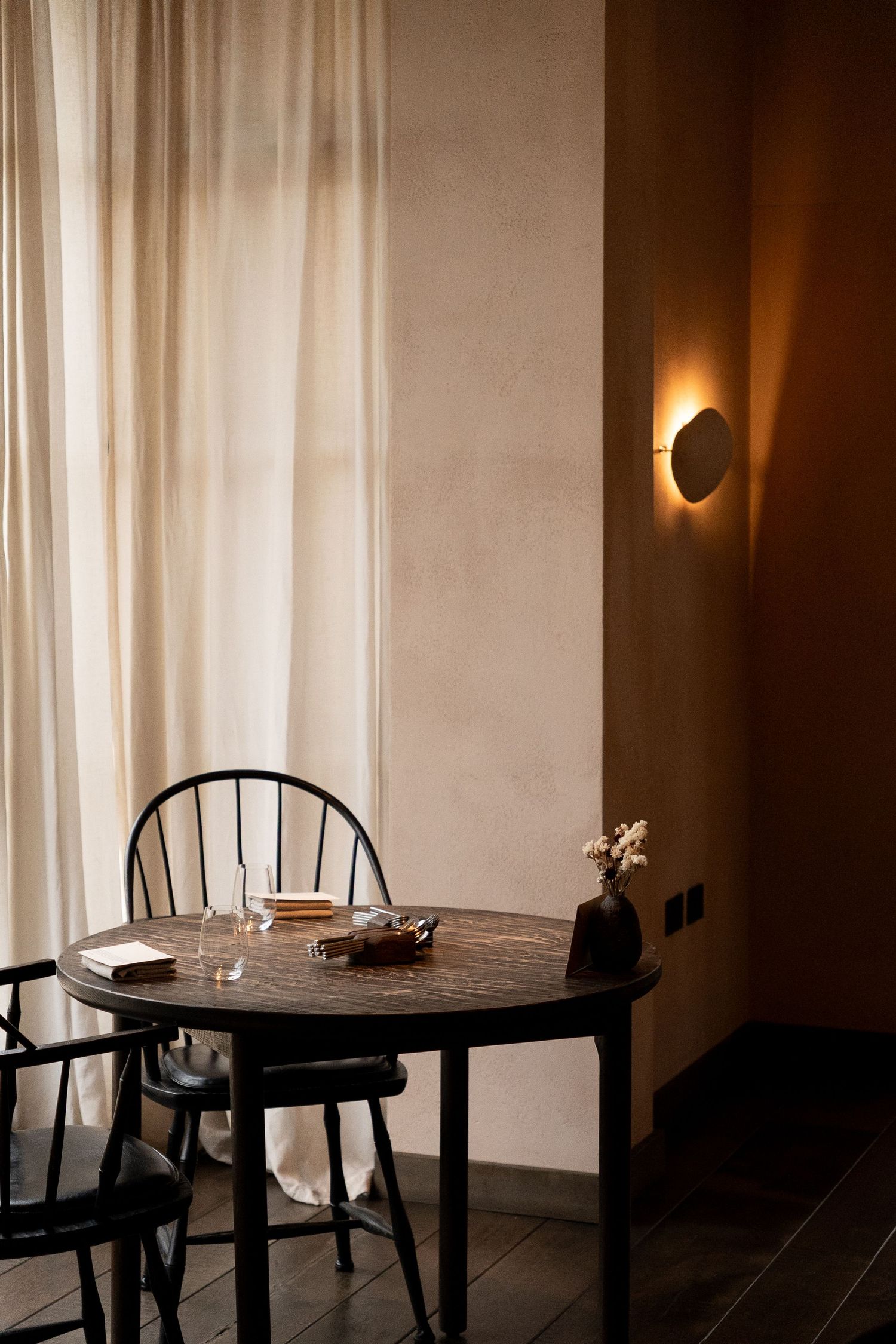 Intimate restaurant corner with dark round table, Windsor chairs, sheer curtains, and warm circular wall sconce lighting.