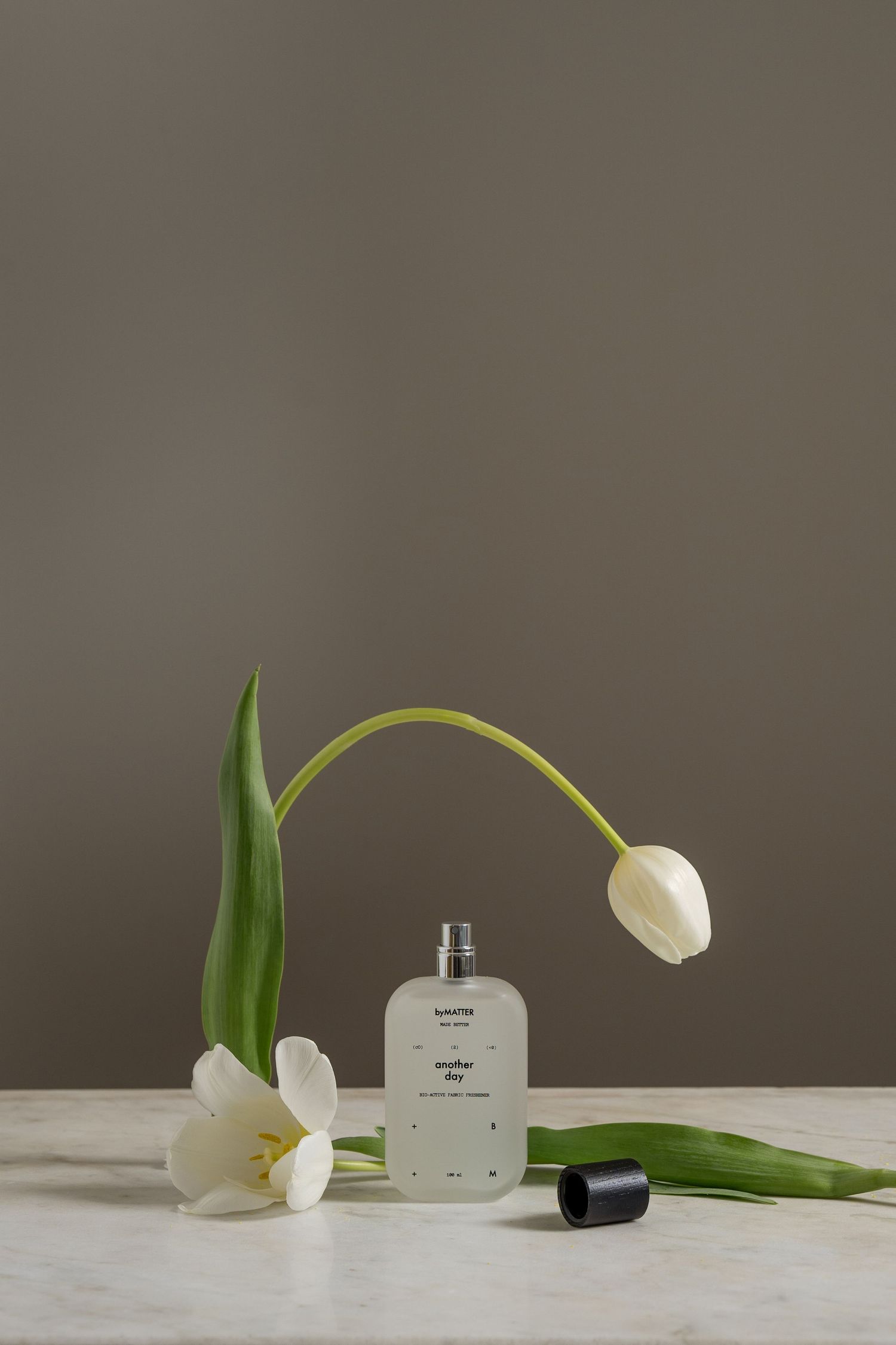 Minimalist perfume bottle named Another Day by Luzette displayed with fresh white tulips on marble surface.