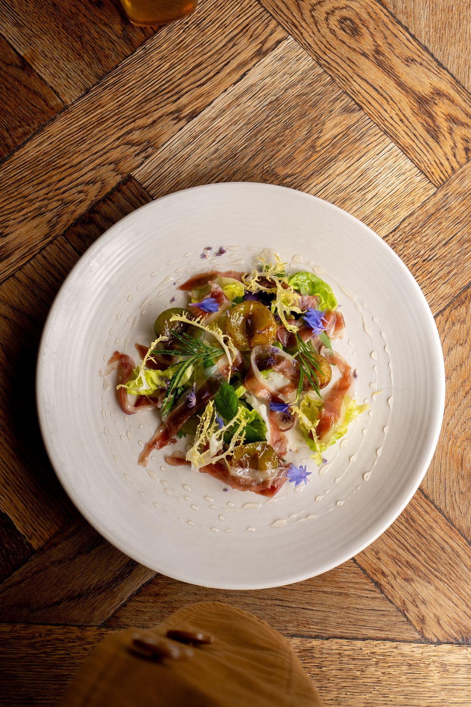 An elegant restaurant salad with edible flowers, fresh greens, and sauce on a white plate atop a herringbone wood table.