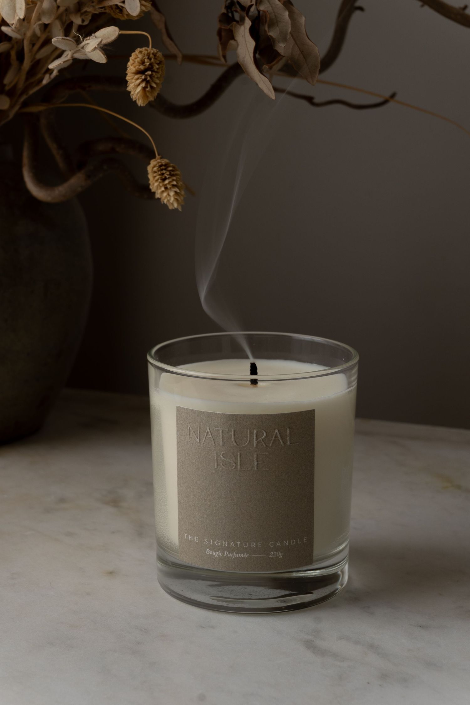 A lit Natural Gel signature candle in a glass jar with smoke rising, beside a dried floral arrangement.