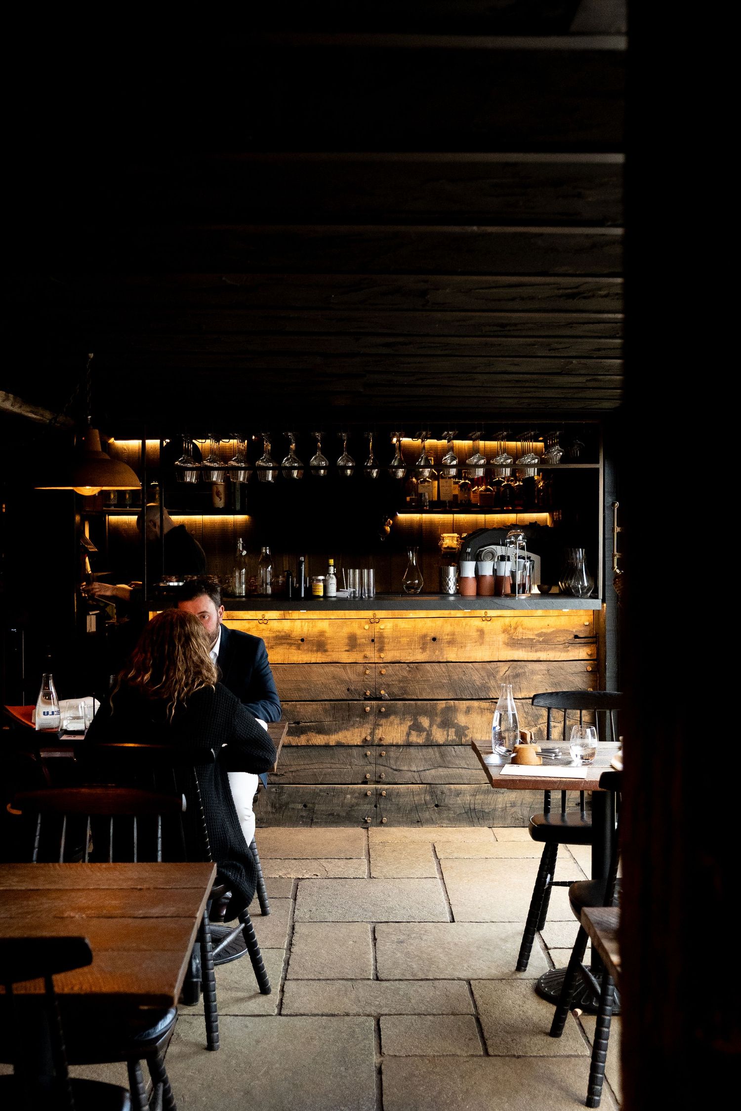 Dimly lit rustic restaurant interior with wooden bar and patron seated at table.