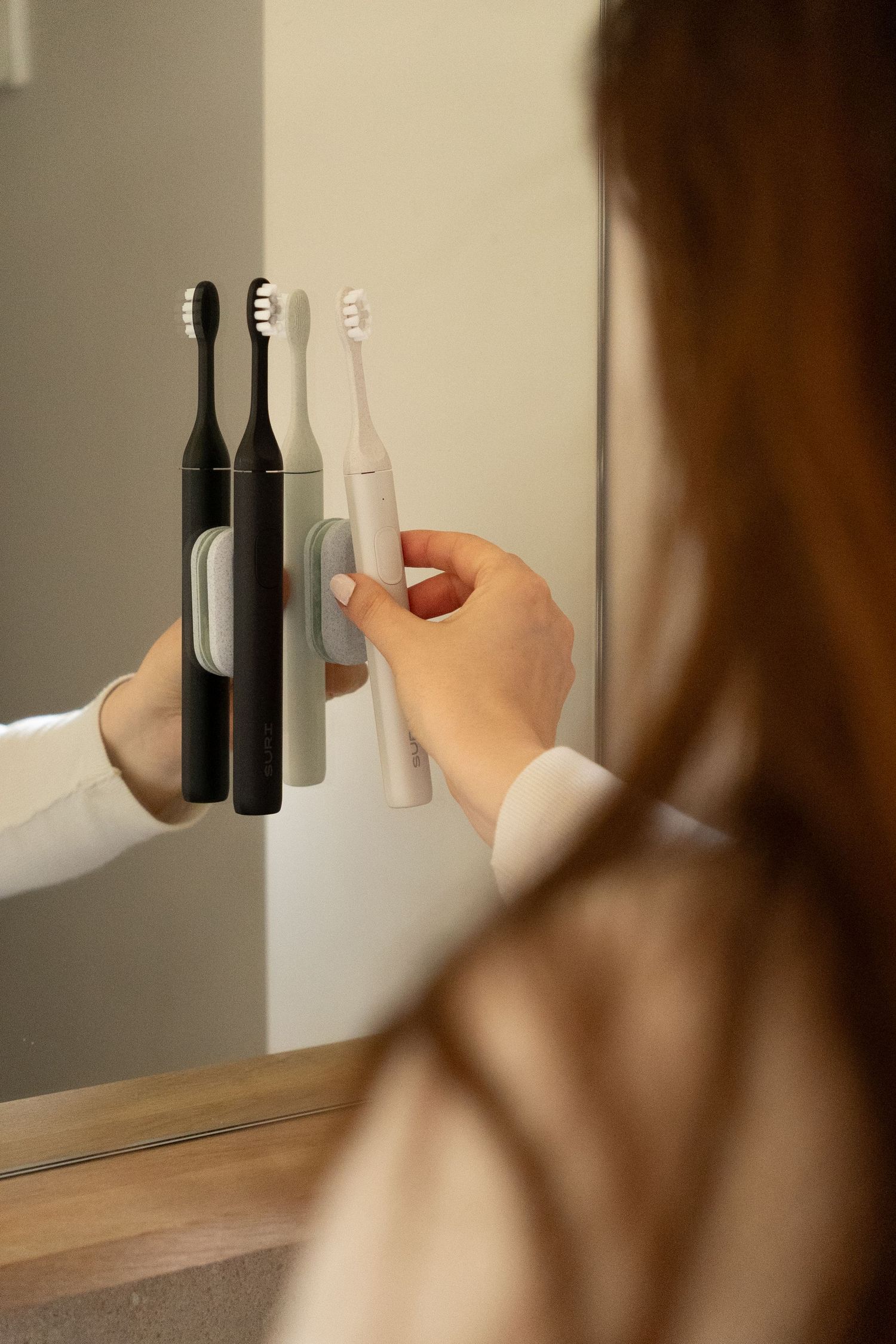 Woman with red hair placing a white electric toothbrush into a mirror-mounted three-slot holder.