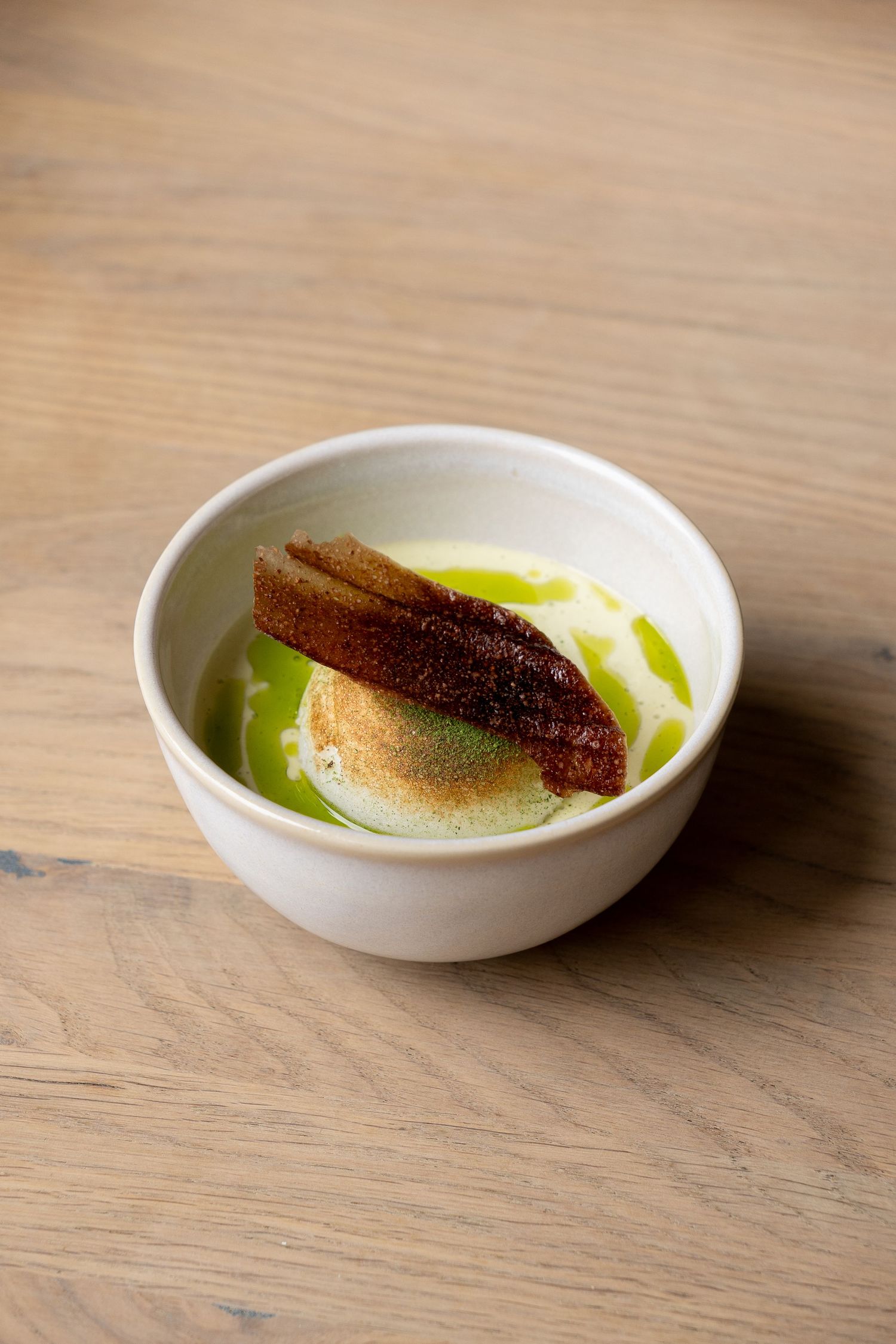 A white ceramic bowl with green herb oil, creamy puree, and crispy fish skin on a wooden table.