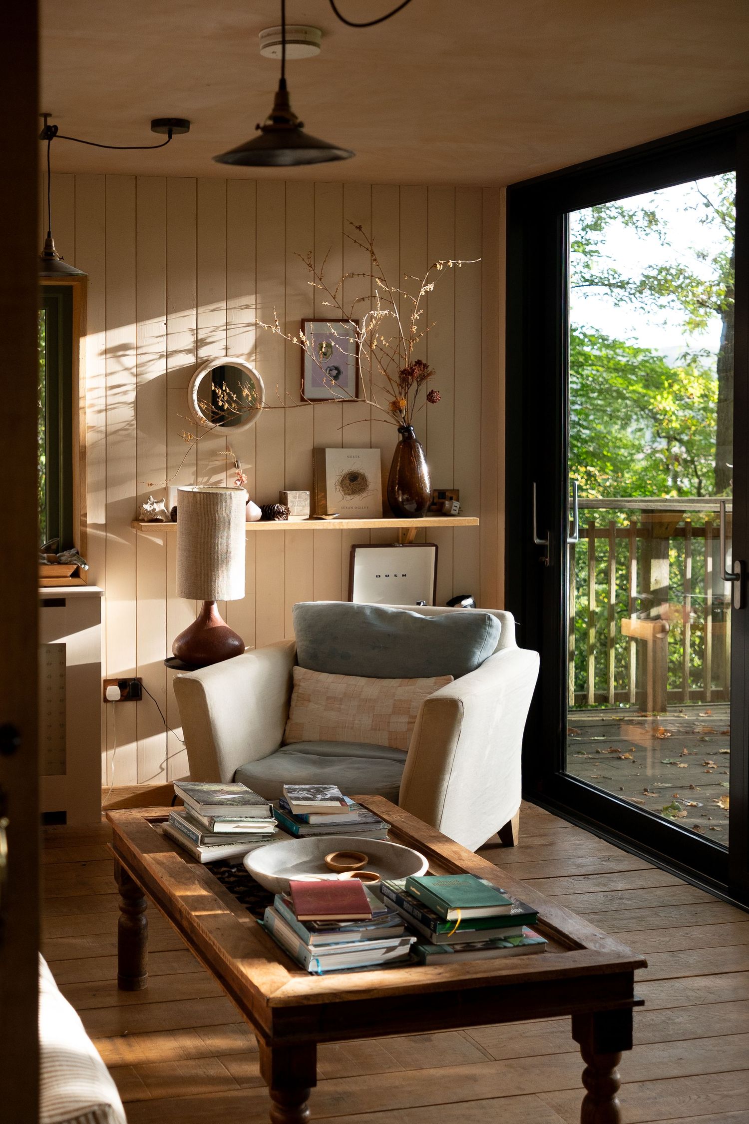 Cozy cabin living room with white armchair, rustic wood coffee table stacked with books, and large glass door to forest deck.