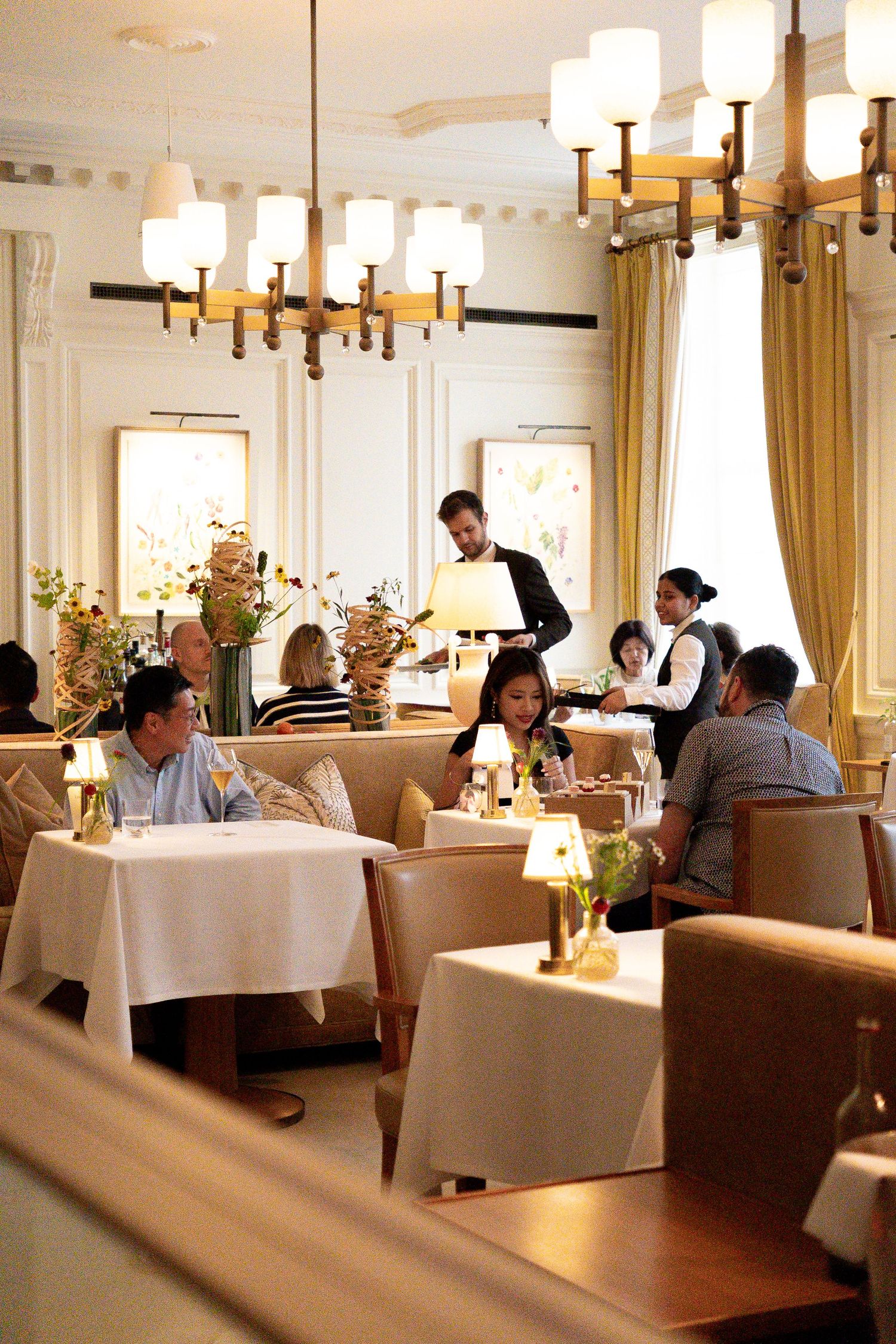 Busy upscale restaurant dining room with chandeliers, gold drapes, and staff serving seated guests.