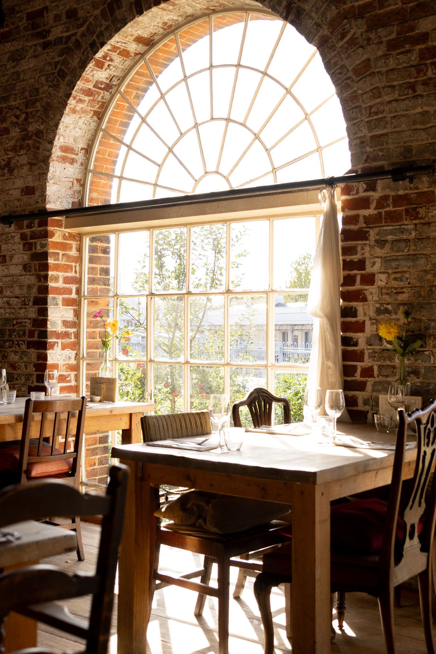 Sunlit rustic restaurant interior with arched brick window, wooden tables, and wine glasses in warm natural light.