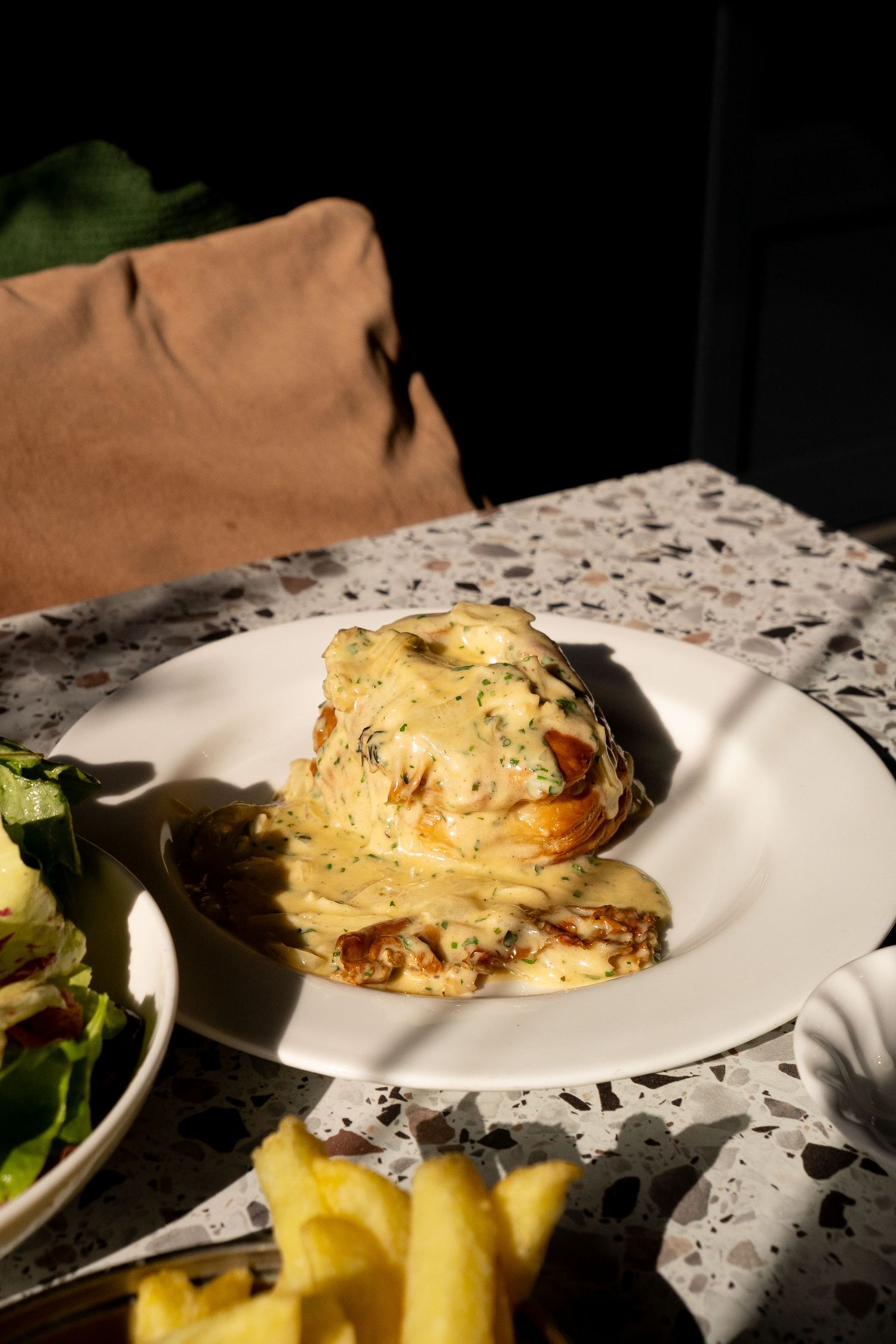Cheesy baked dish with golden sauce served on a white plate alongside fries and salad on a terrazzo table.