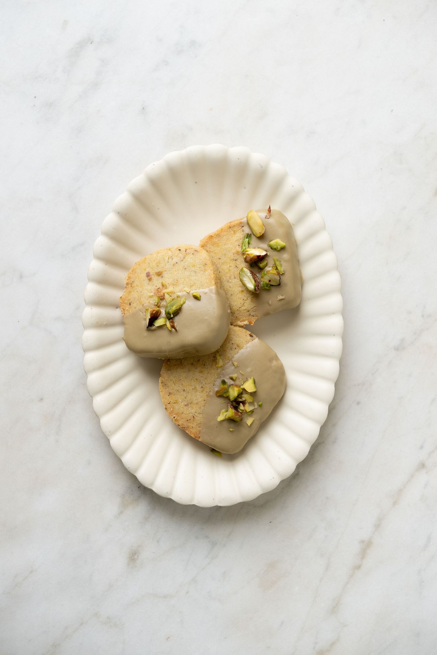 Three pistachio financier cakes with caramel glaze and chopped pistachios on a white fluted oval plate.