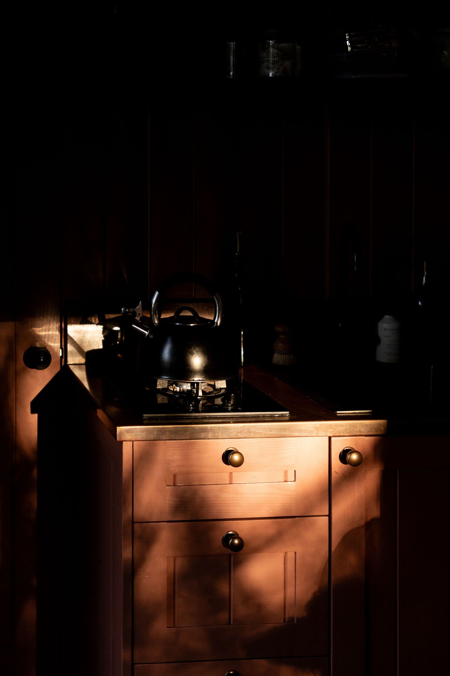 Dramatic moody kitchen with black kettle on stove illuminated by single shaft of warm sunlight.