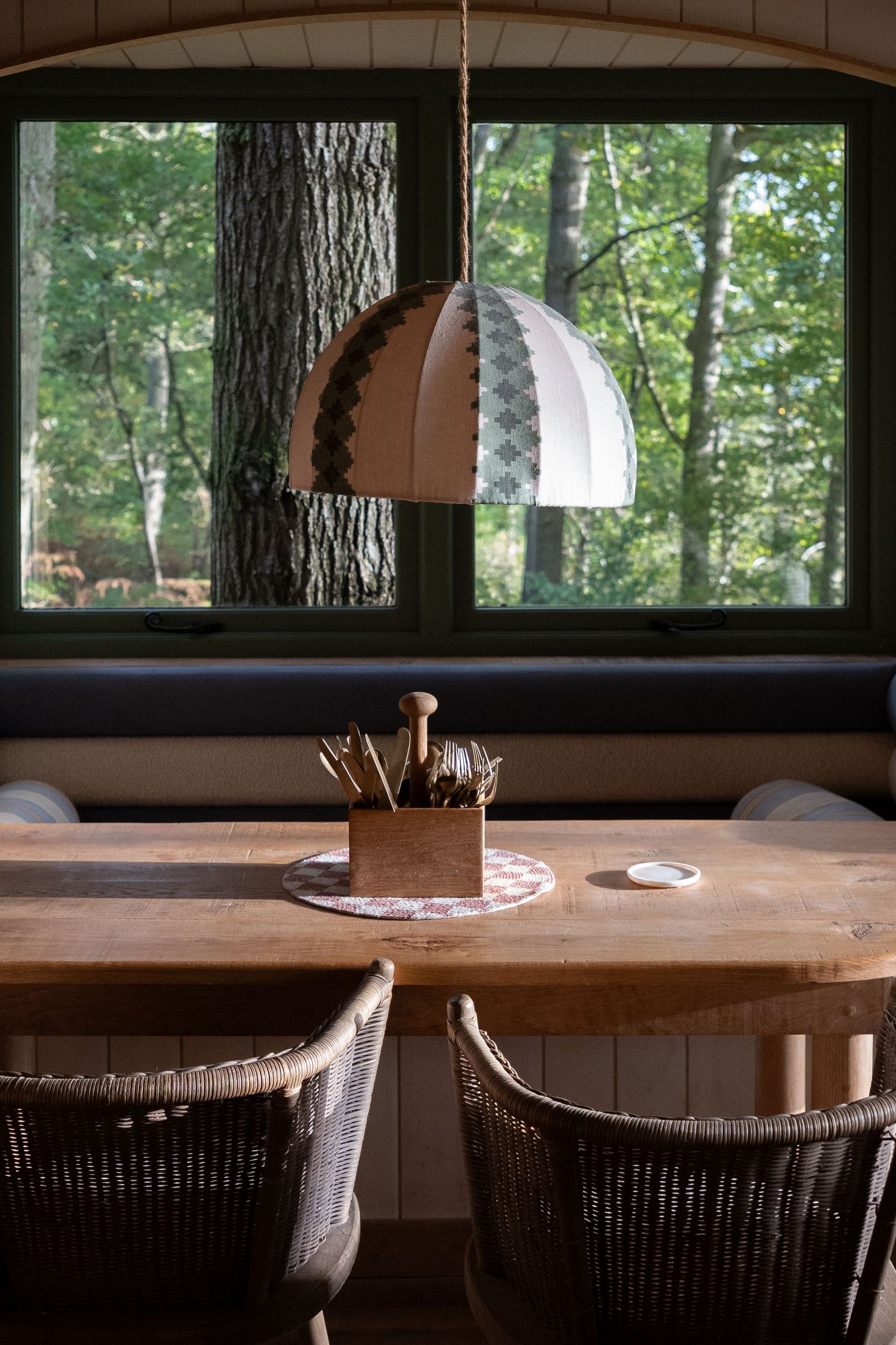Rustic wooden dining table with wicker chairs beneath a dome pendant light, framed by large window overlooking forest.