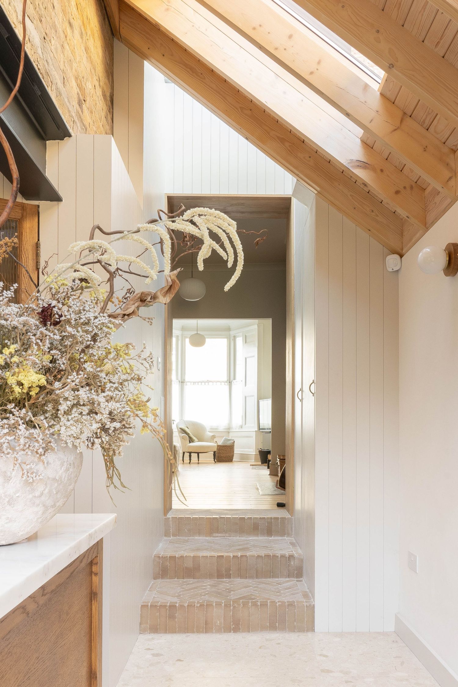 Bright hallway with exposed timber beams, herringbone tile steps, white shiplap walls, and dried floral arrangement.
