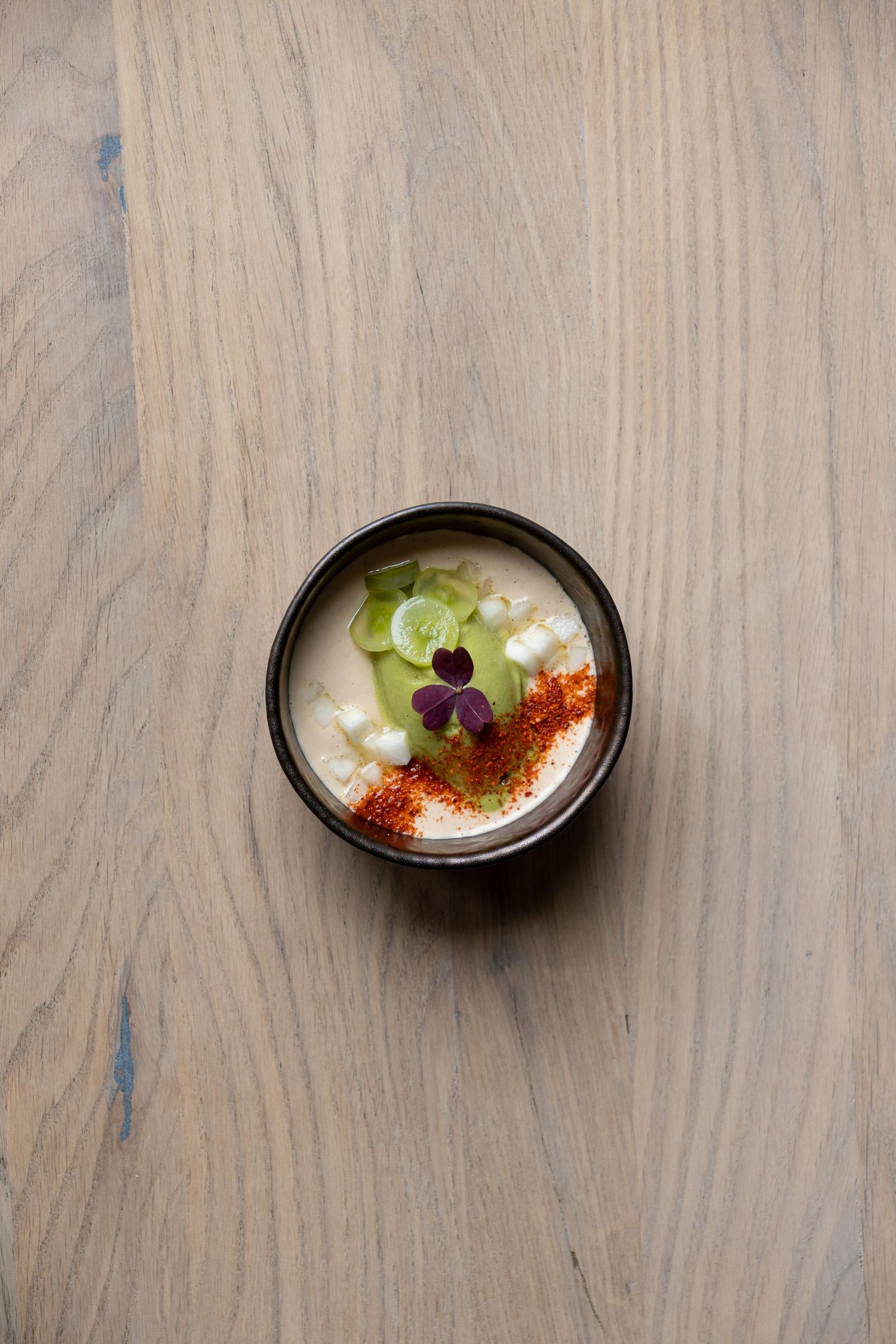 A dark ceramic bowl with creamy white soup, cucumber slices, red spice, and a purple flower garnish.