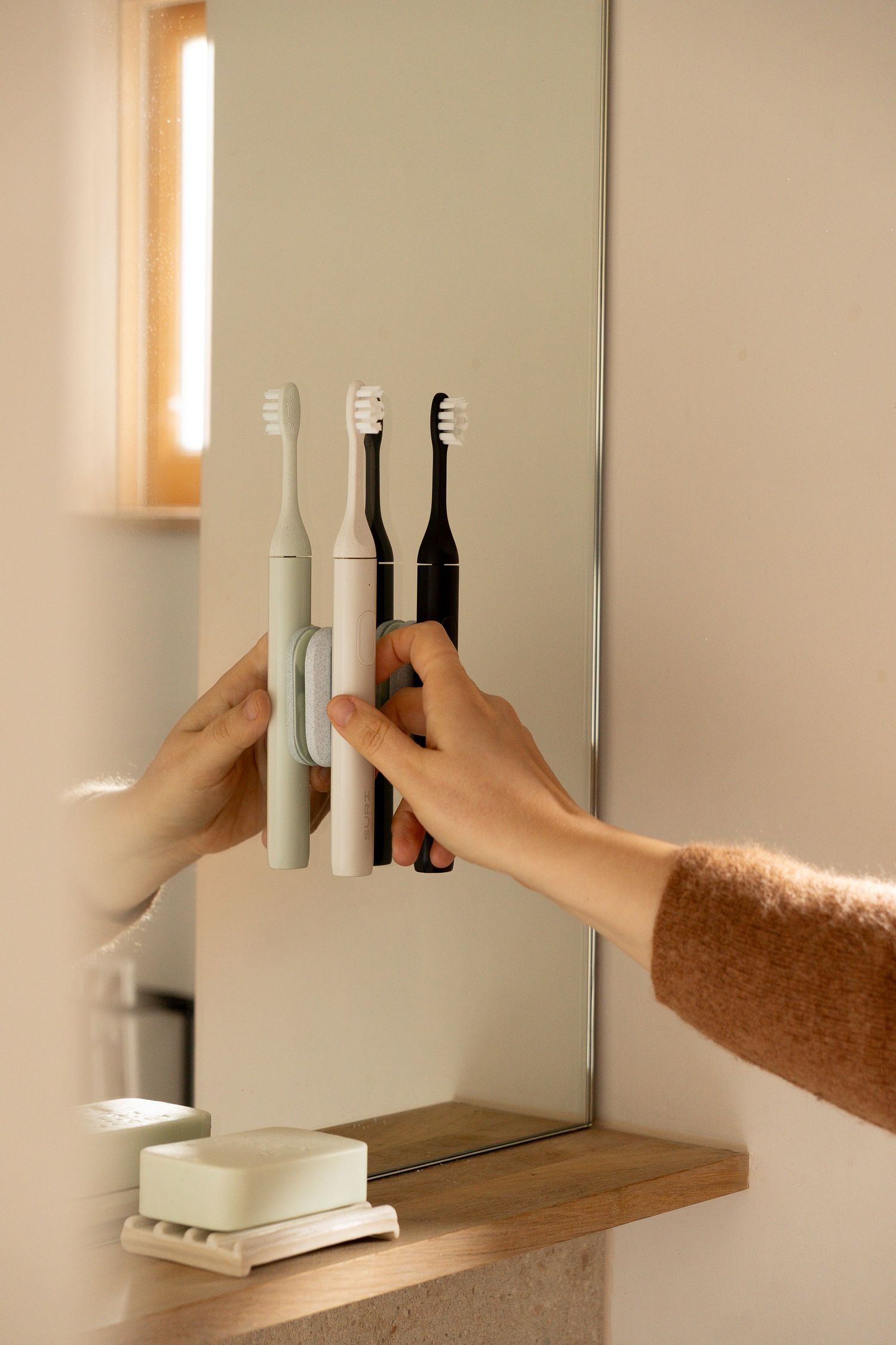 Hands arranging three electric toothbrushes in a wall-mounted holder beside a bathroom mirror.