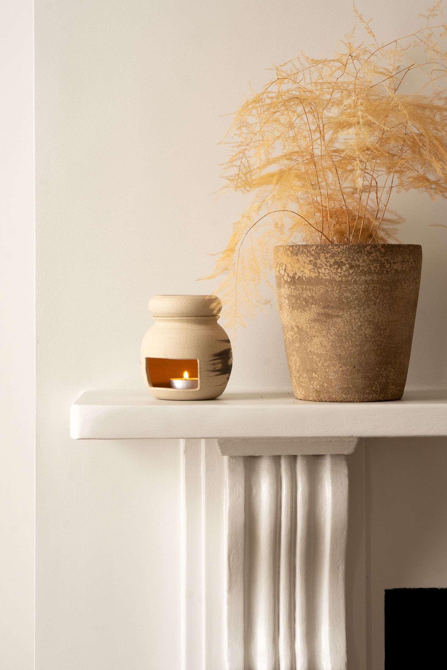 Ceramic oil burner with lit candle beside a terracotta pot with dried grasses on white mantle.