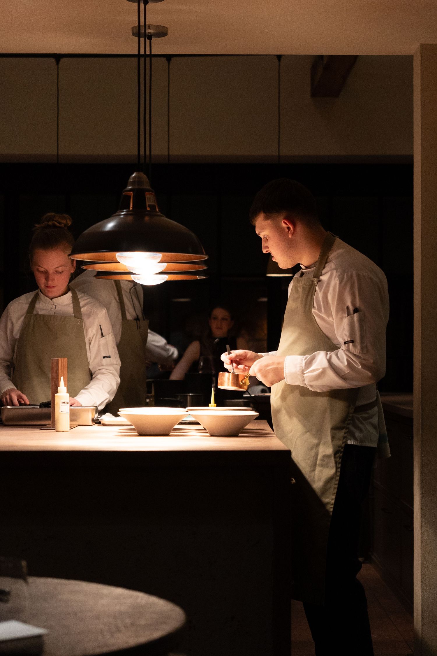 Two chefs in sage green aprons plating dishes under pendant lights in a dimly lit fine dining kitchen.