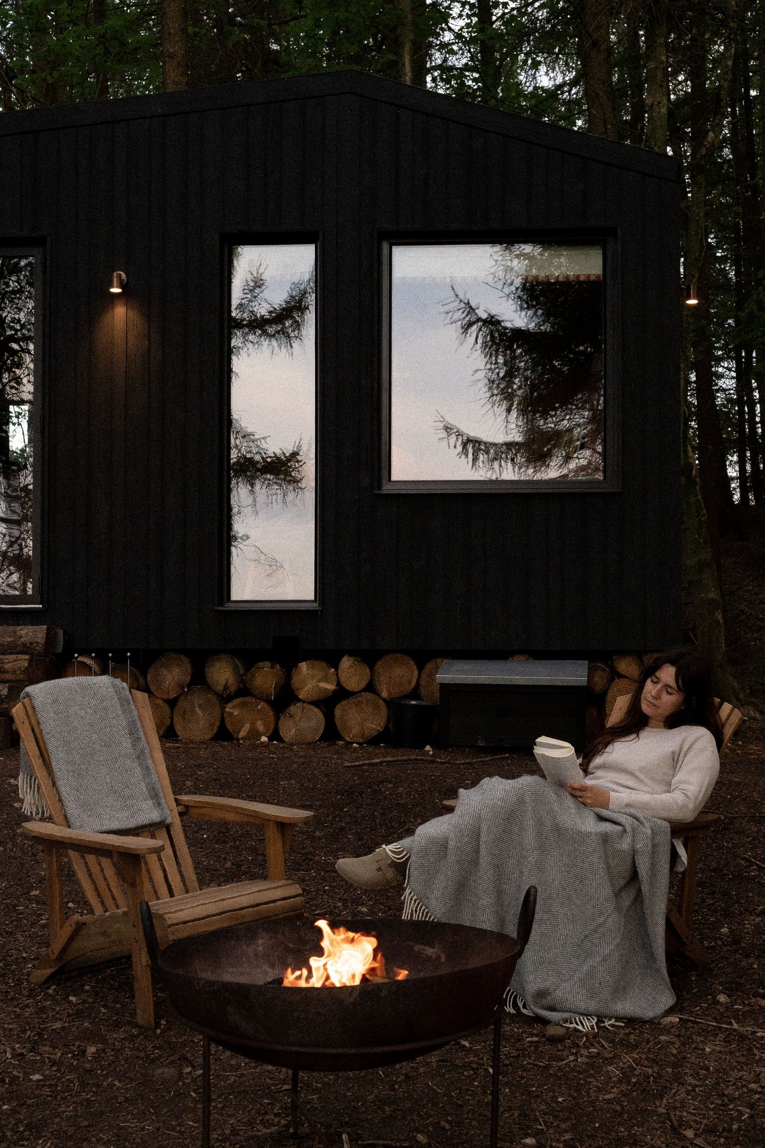 Woman reading a book wrapped in blanket by a fire pit outside a modern black tiny cabin in the woods.