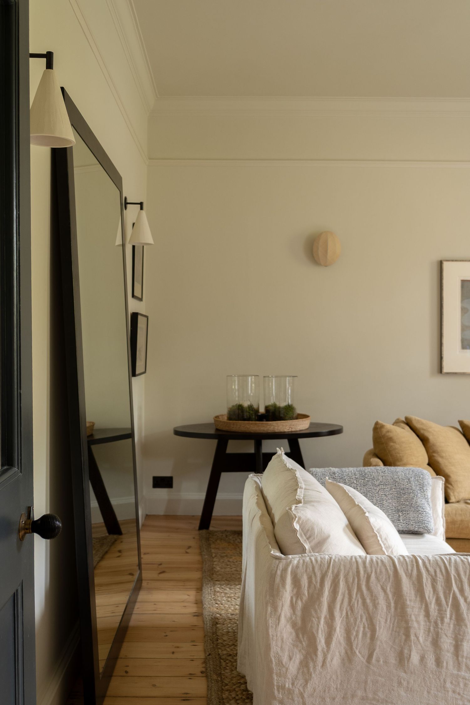 Minimalist living room with linen sofa, floor mirror, wall sconce, and round dark wood accent table.