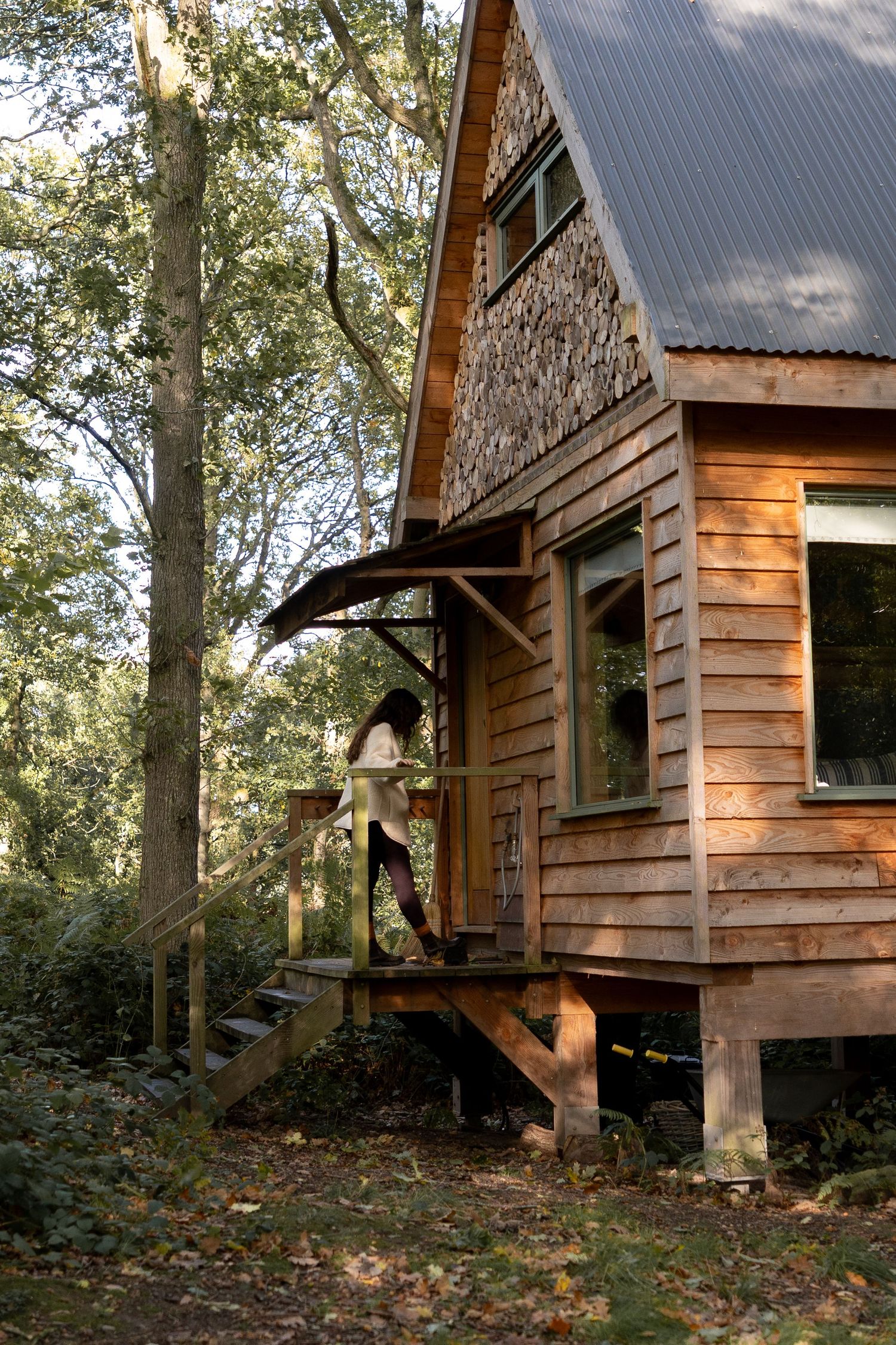 Woman standing on wooden steps of rustic A-frame cabin with cedar siding and metal roof surrounded by forest trees.
