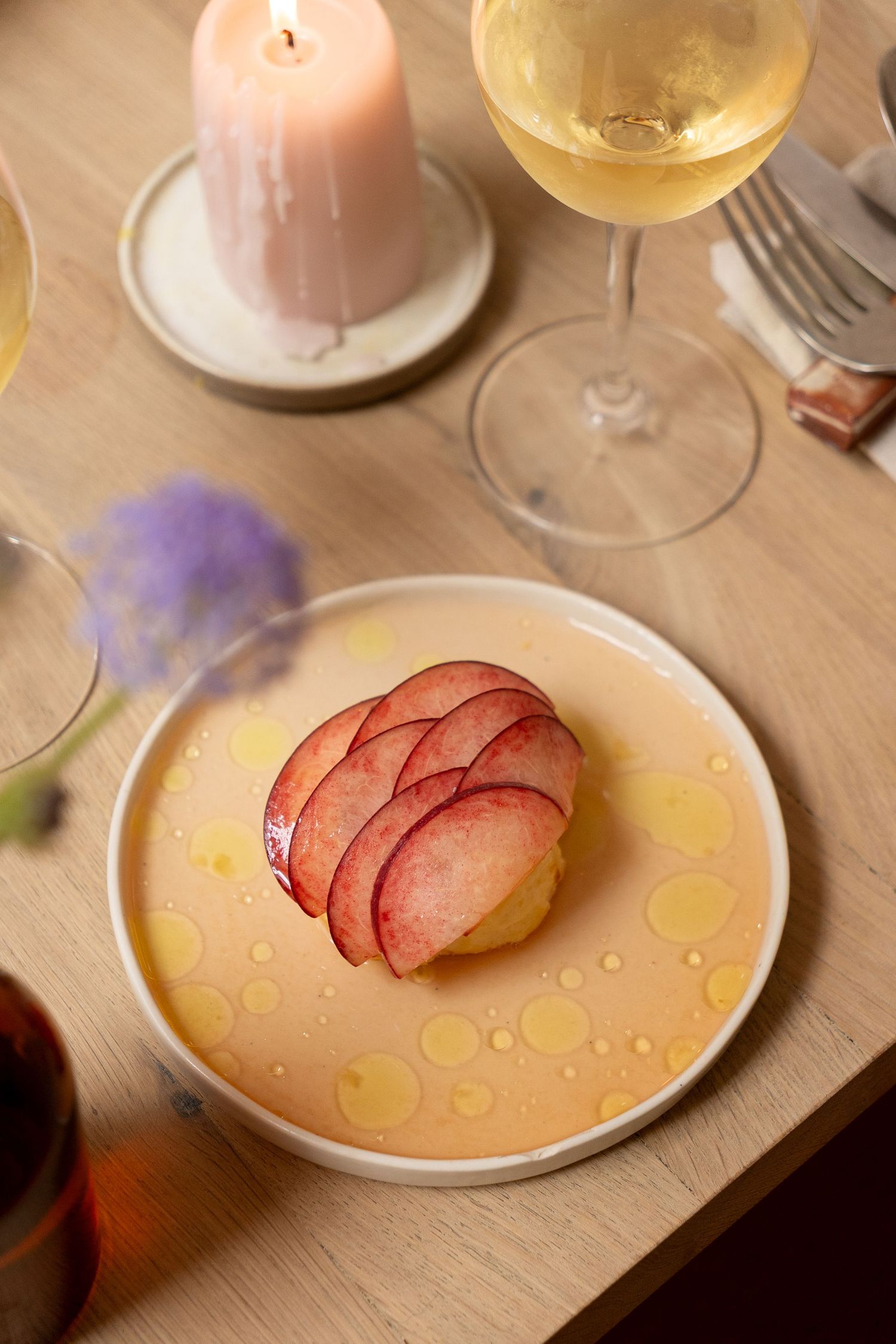 Elegant dessert plate with thinly sliced red apple fan on cream sauce, beside a lit candle and white wine glass.
