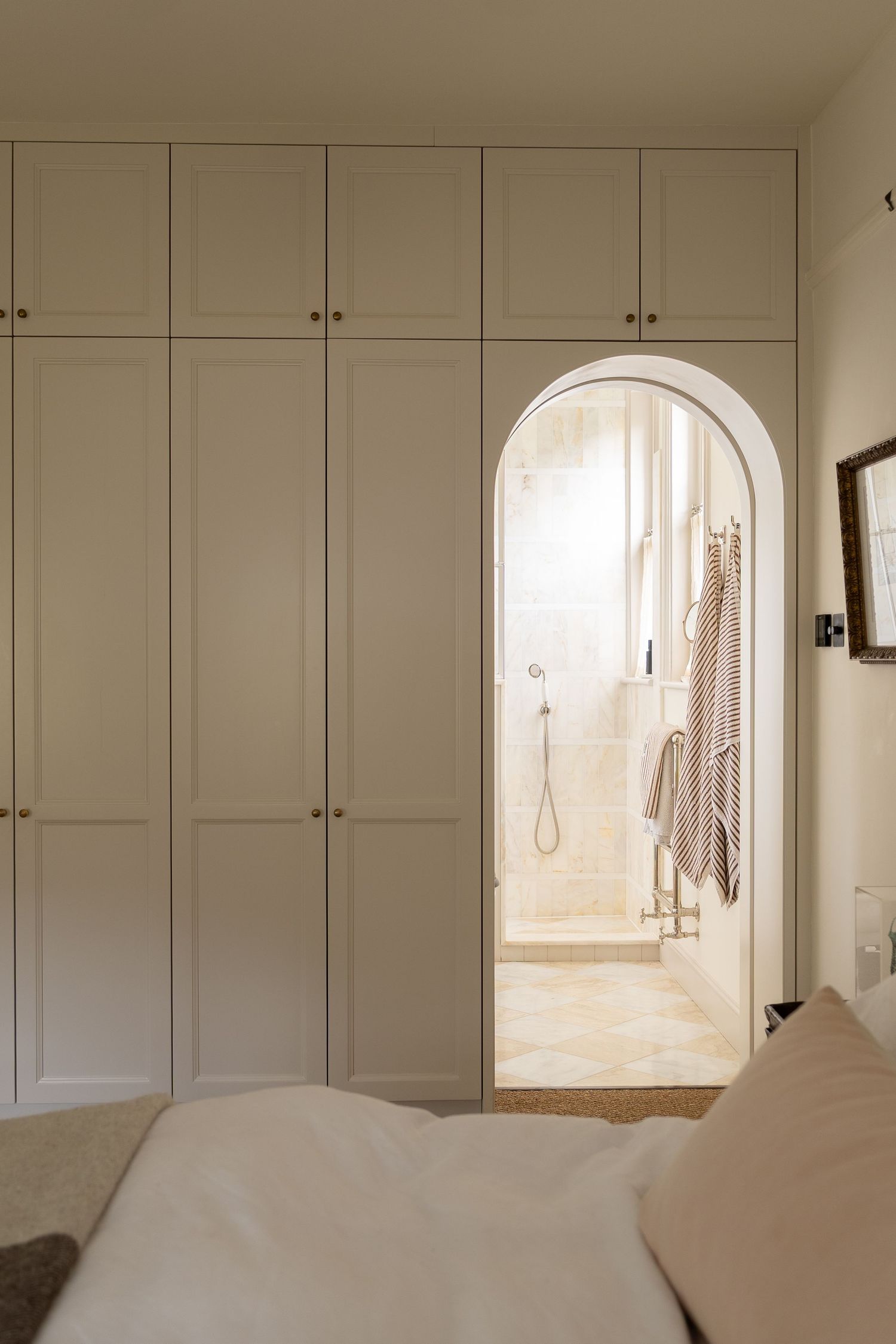 Built-in cream wardrobe wall with arched doorway leading to marble ensuite bathroom in elegant bedroom.