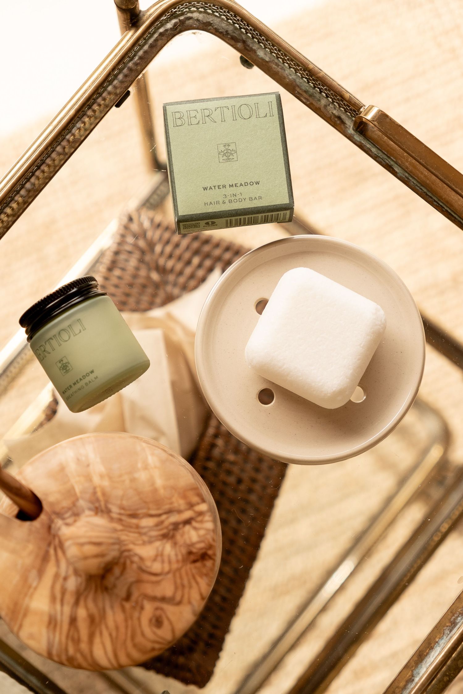 Overhead view of brass tray with Bertioli soap bar, small candle jar, and wooden accessories on wicker surface.