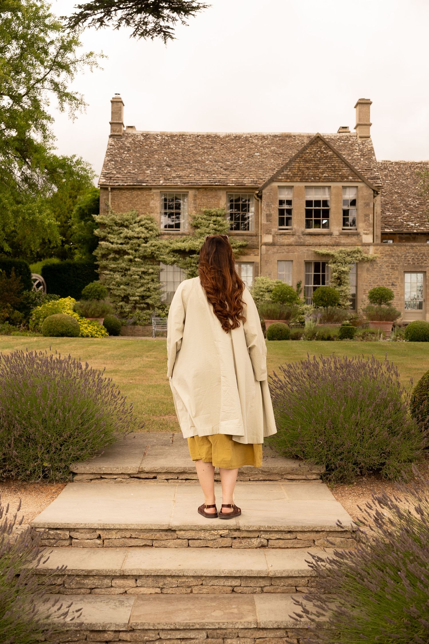 Woman in cream coat standing before a grand Cotswolds stone manor house with lavender gardens.