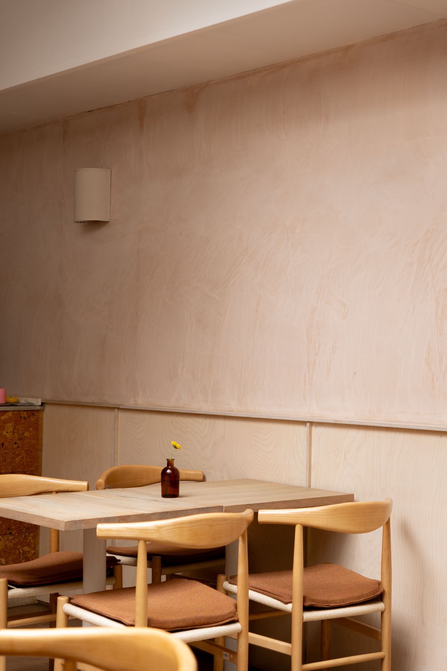 Minimalist cafe interior with light wood chairs, concrete table, and a small amber vase with yellow flower against a warm plaster wall.