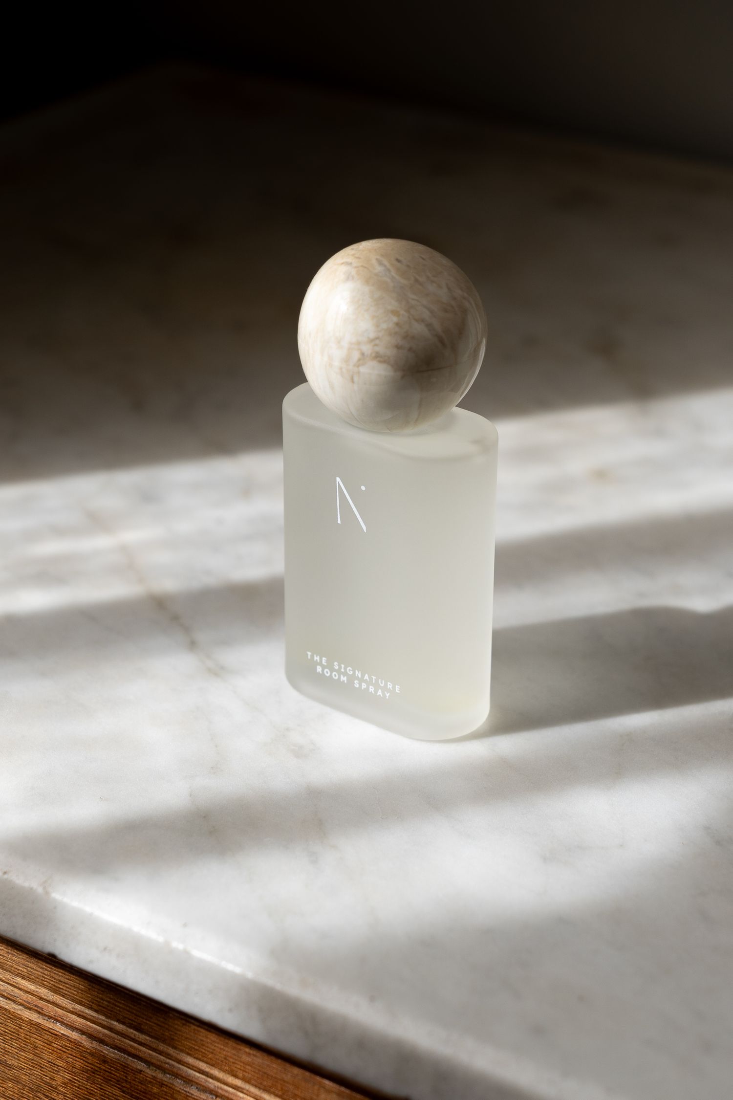 N. Signature Room Spray frosted square glass bottle with marble stone cap on white marble surface in dramatic sunlight.