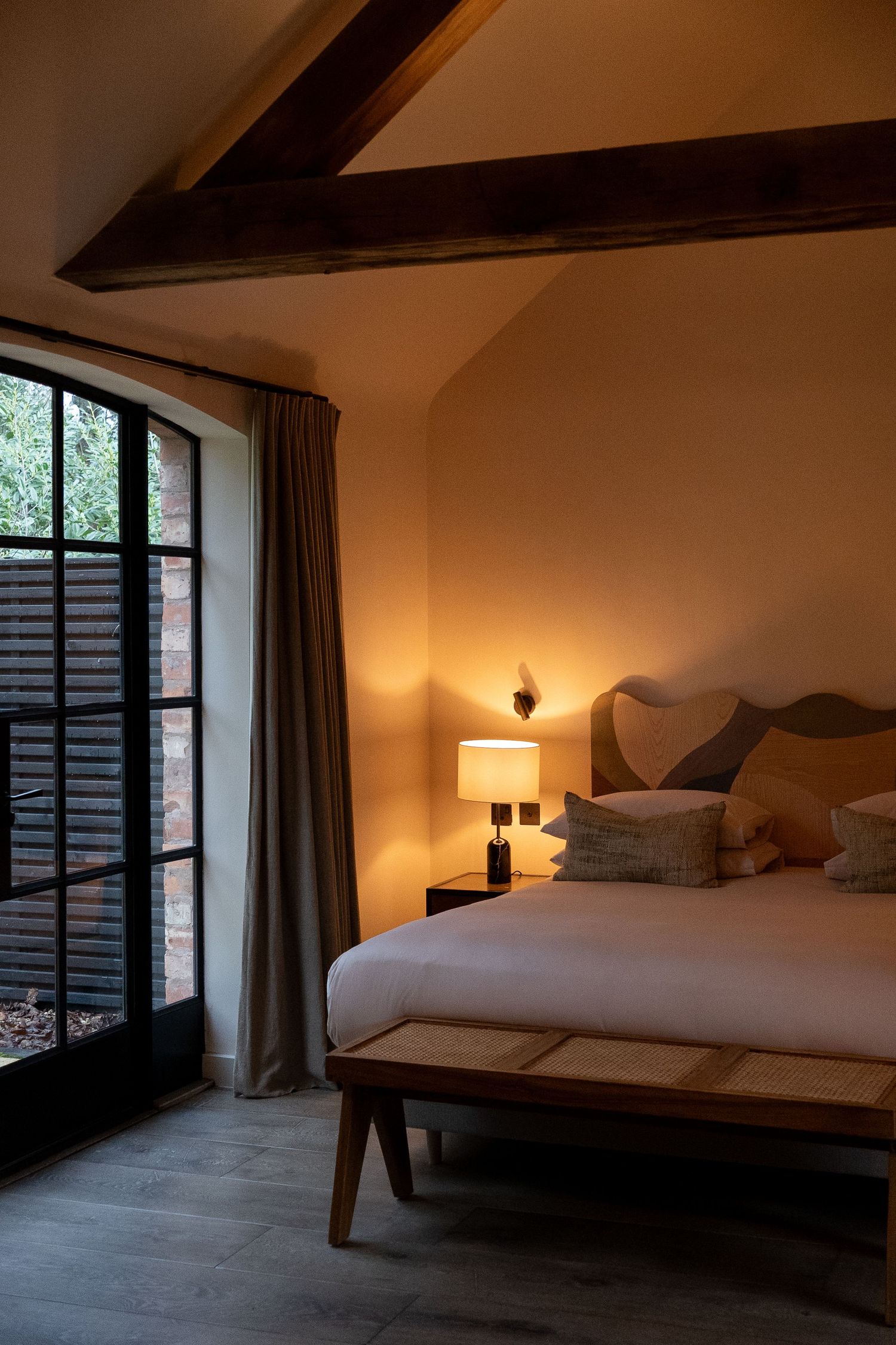 Cozy boutique hotel bedroom with exposed wooden beams, arched steel window, warm bedside lamp, and neutral linen bedding.