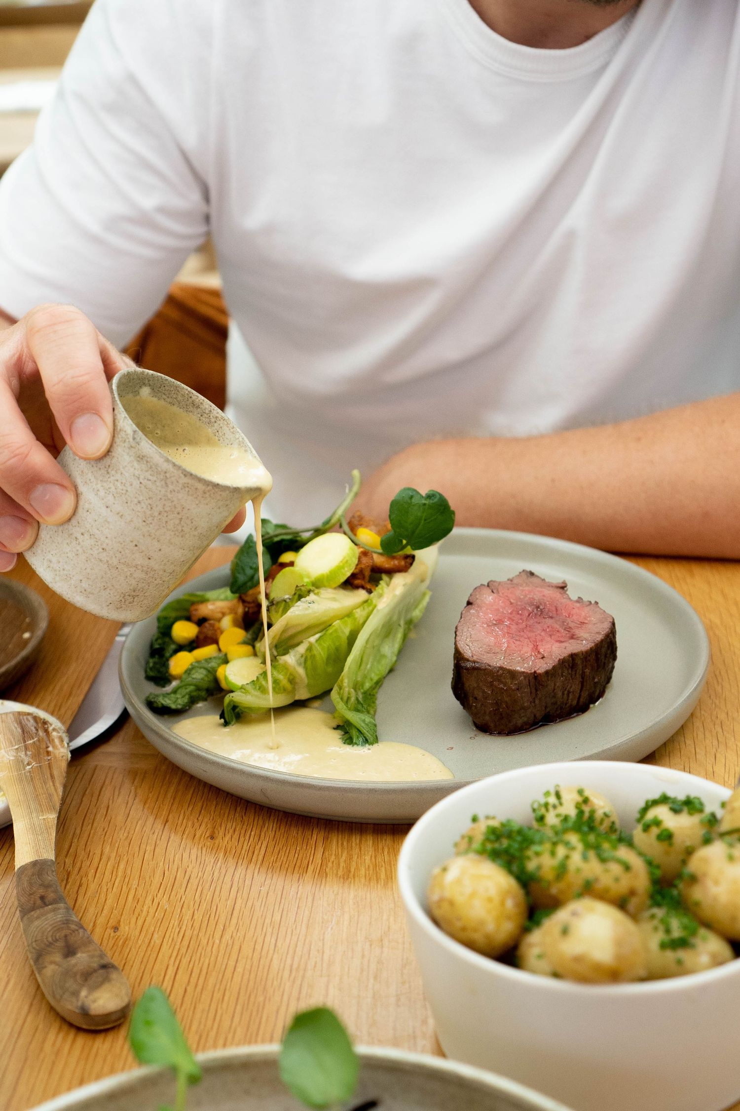 Chef pouring creamy sauce over plated beef fillet with vegetables and new potatoes.