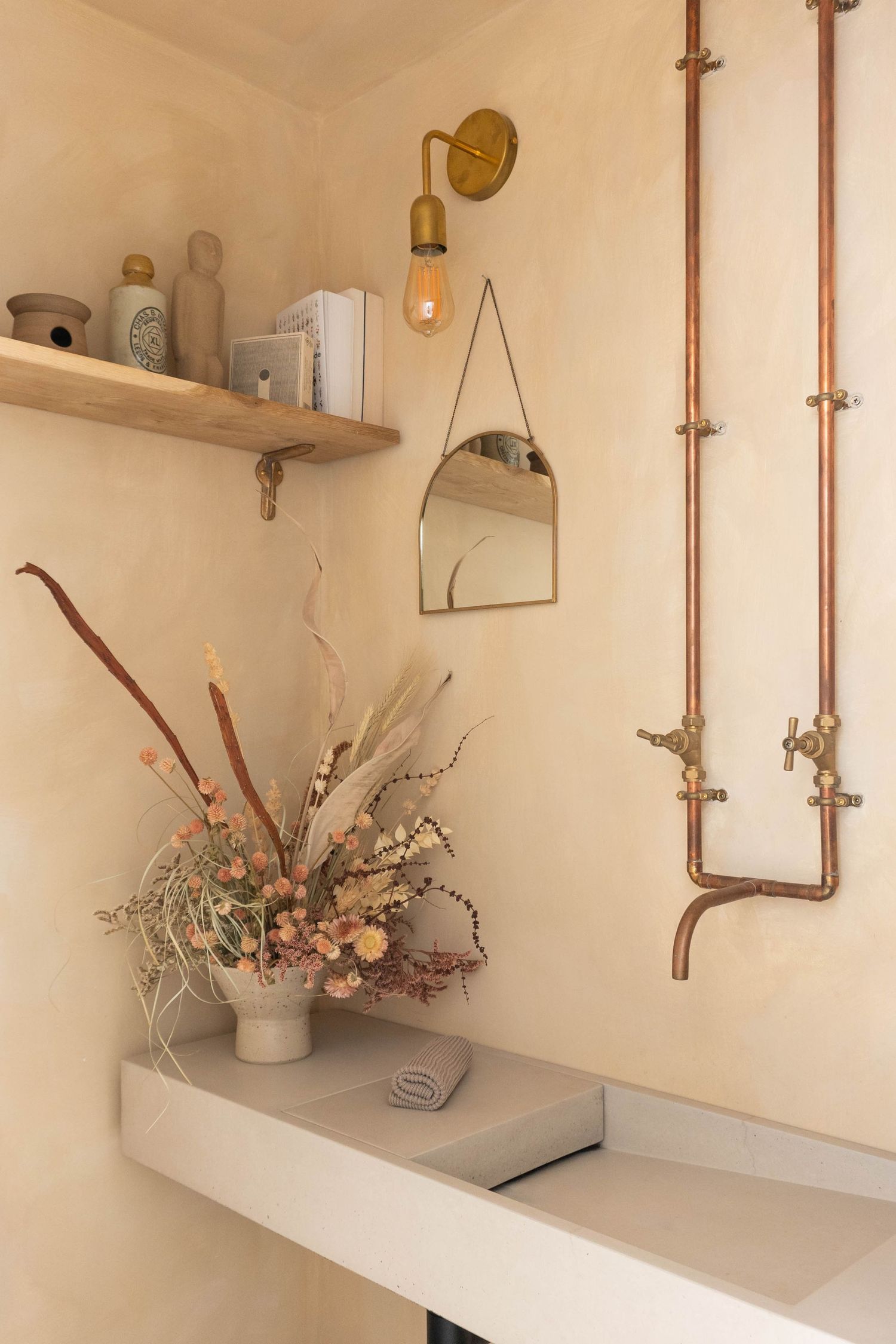 Warm plaster bathroom nook with copper pipe shower, brass sconce, dried floral arrangement, and wood shelf.