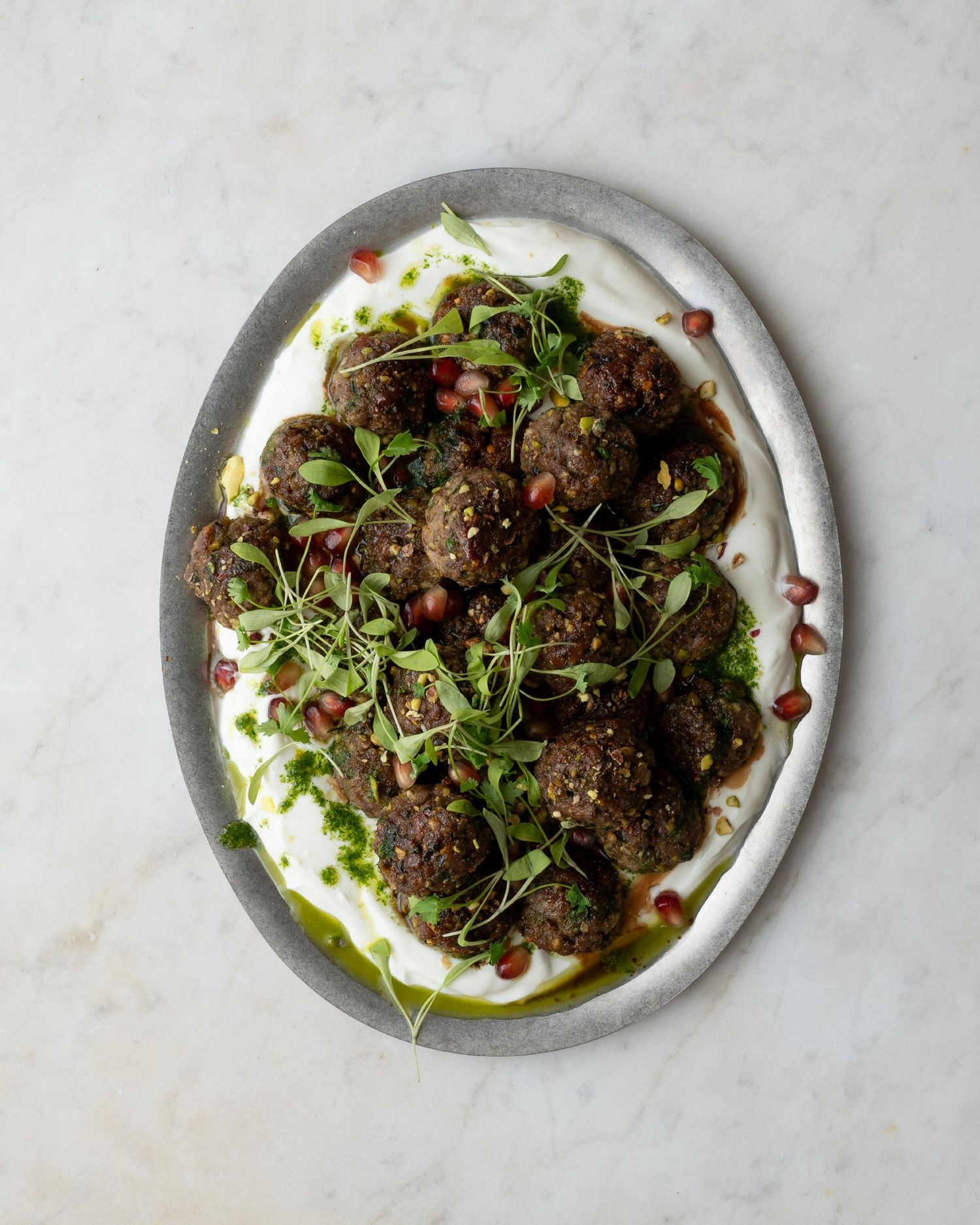 Oval platter of herb-crusted meatballs on white sauce, topped with pomegranate seeds and microgreens.