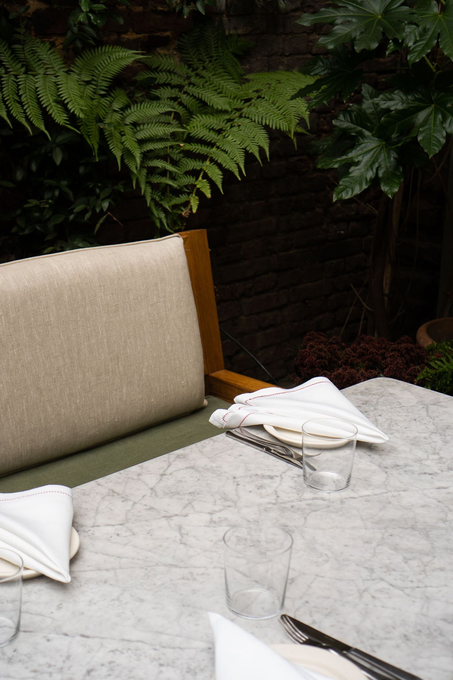 Elegant outdoor restaurant table setting with marble top, white napkins and lush green fern backdrop.