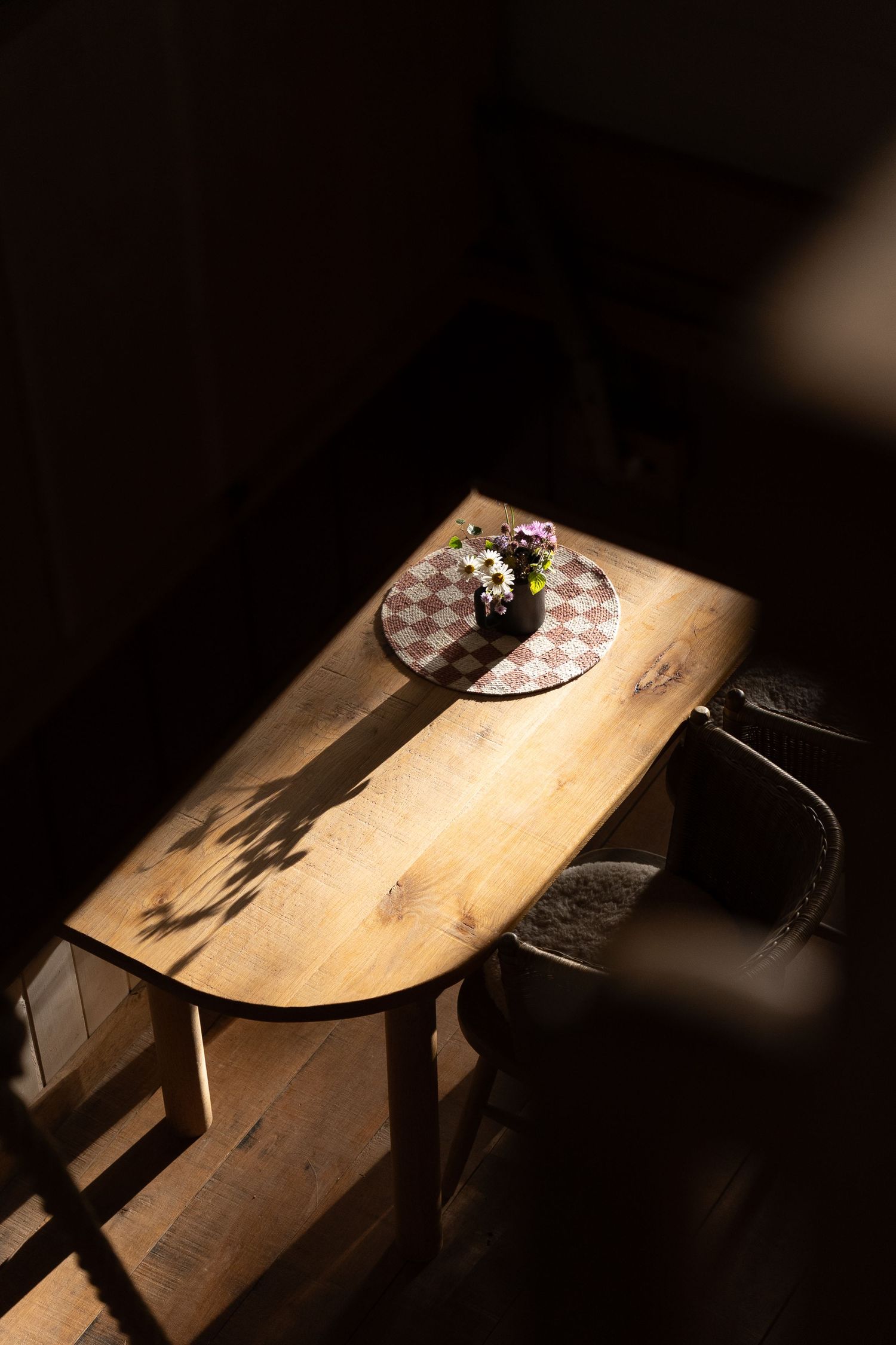 Wooden half-moon table with a checkered placemat and small floral vase illuminated by dramatic sunlight.