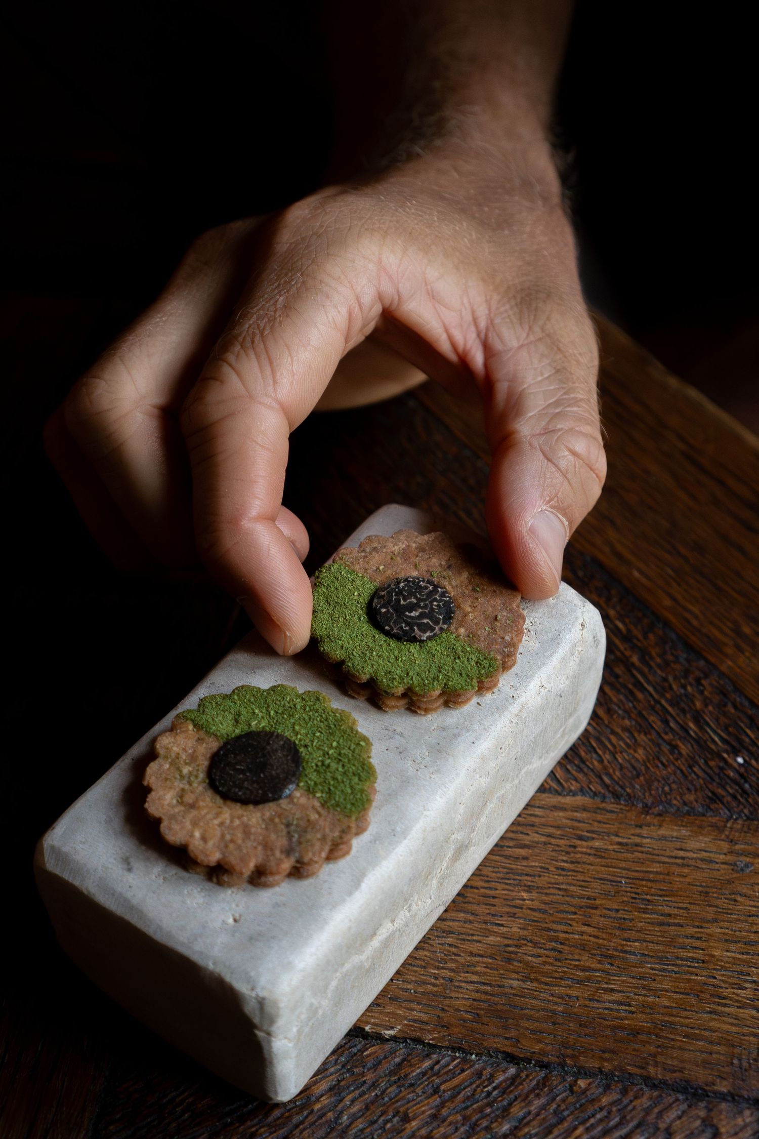 Hand picking up a decorative flower-shaped cookie with matcha green tea powder on a white marble block.