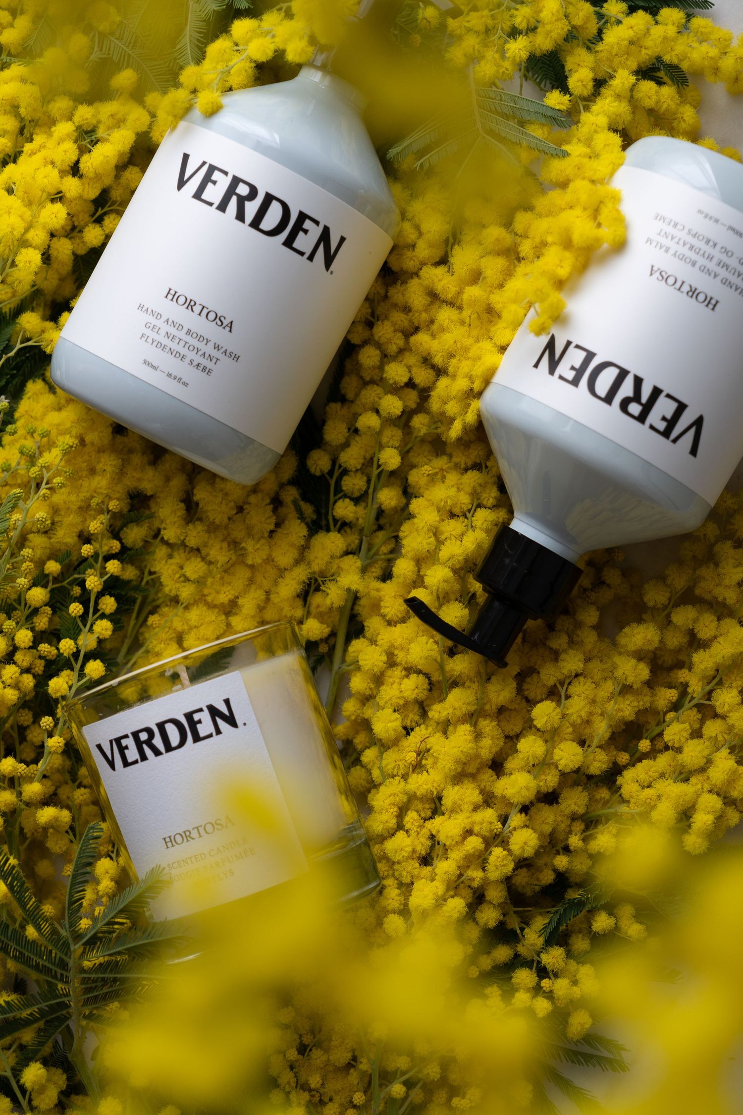 Verden Hortosa hand wash, lotion, and candle set displayed among vibrant yellow mimosa flowers.