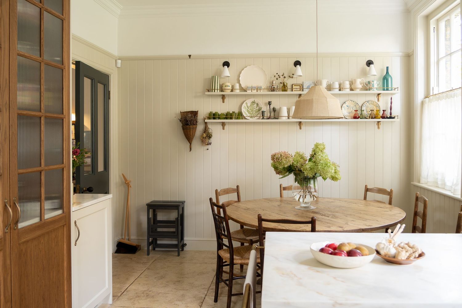 Bright farmhouse kitchen with round wooden dining table, mismatched chairs, and open display shelving.