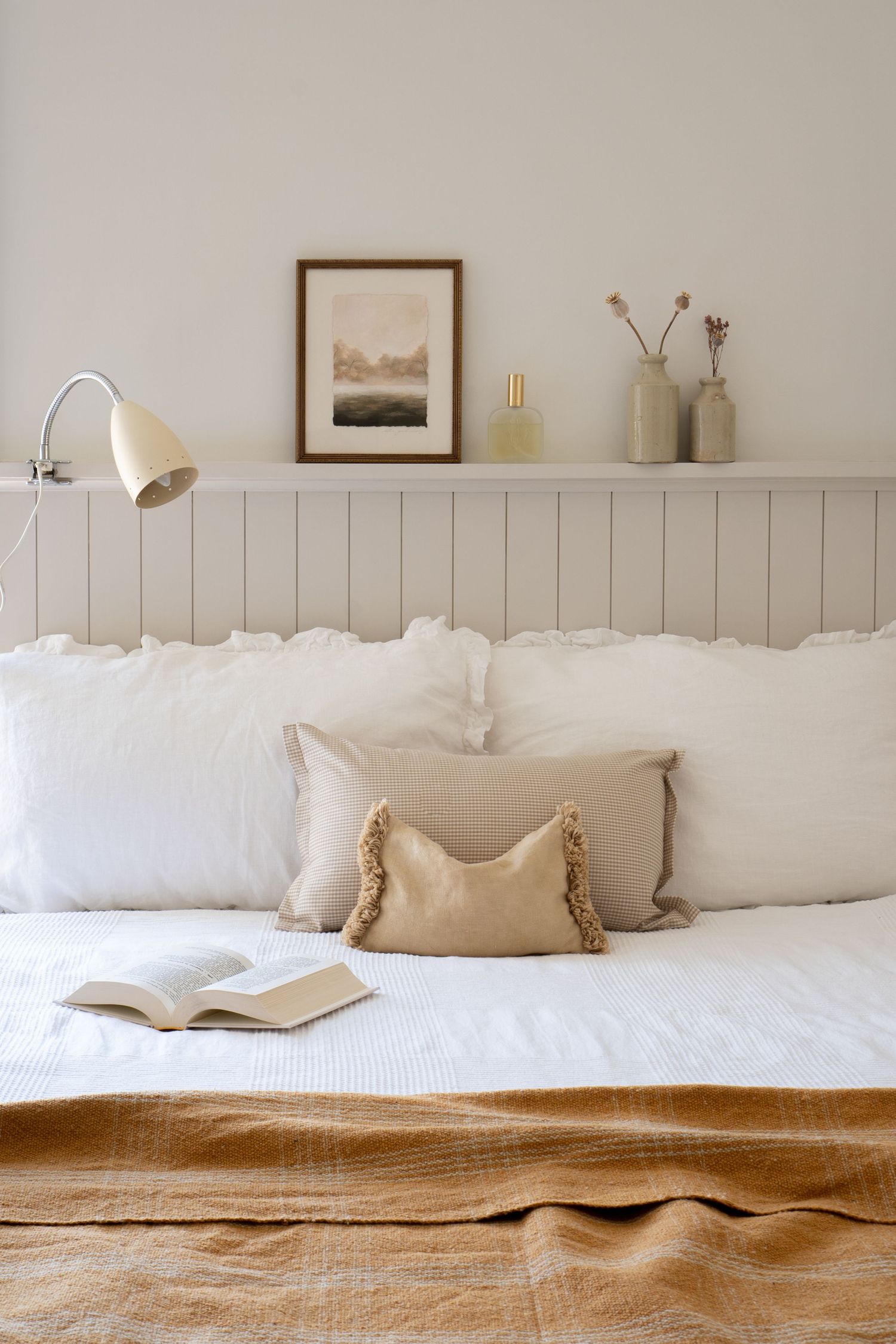 Serene bedroom with paneled headboard, layered neutral pillows, open book, mustard blanket, and decorative shelf above.