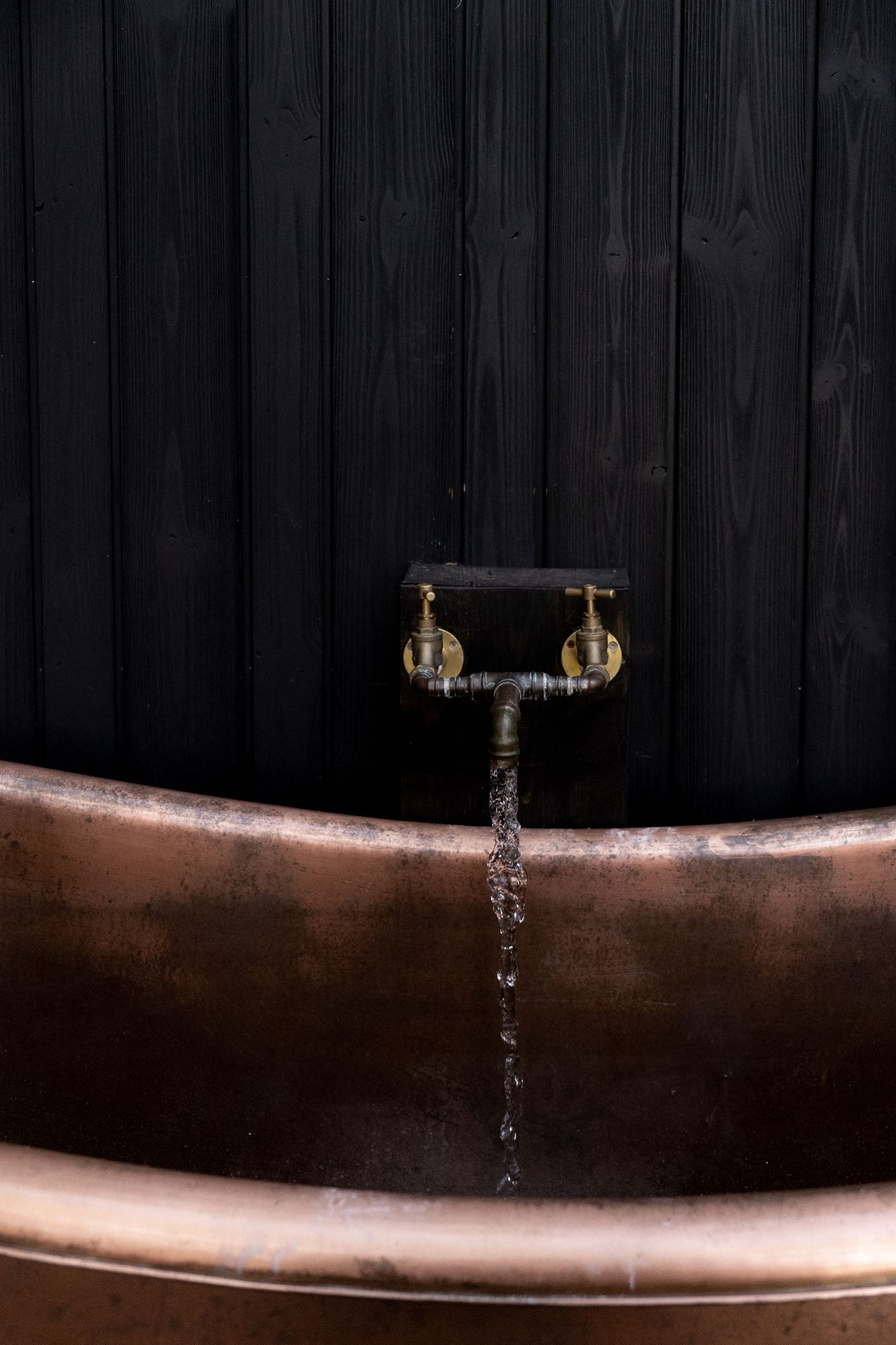 Vintage copper bathtub with antique brass globe faucet running water against dark black wood wall.