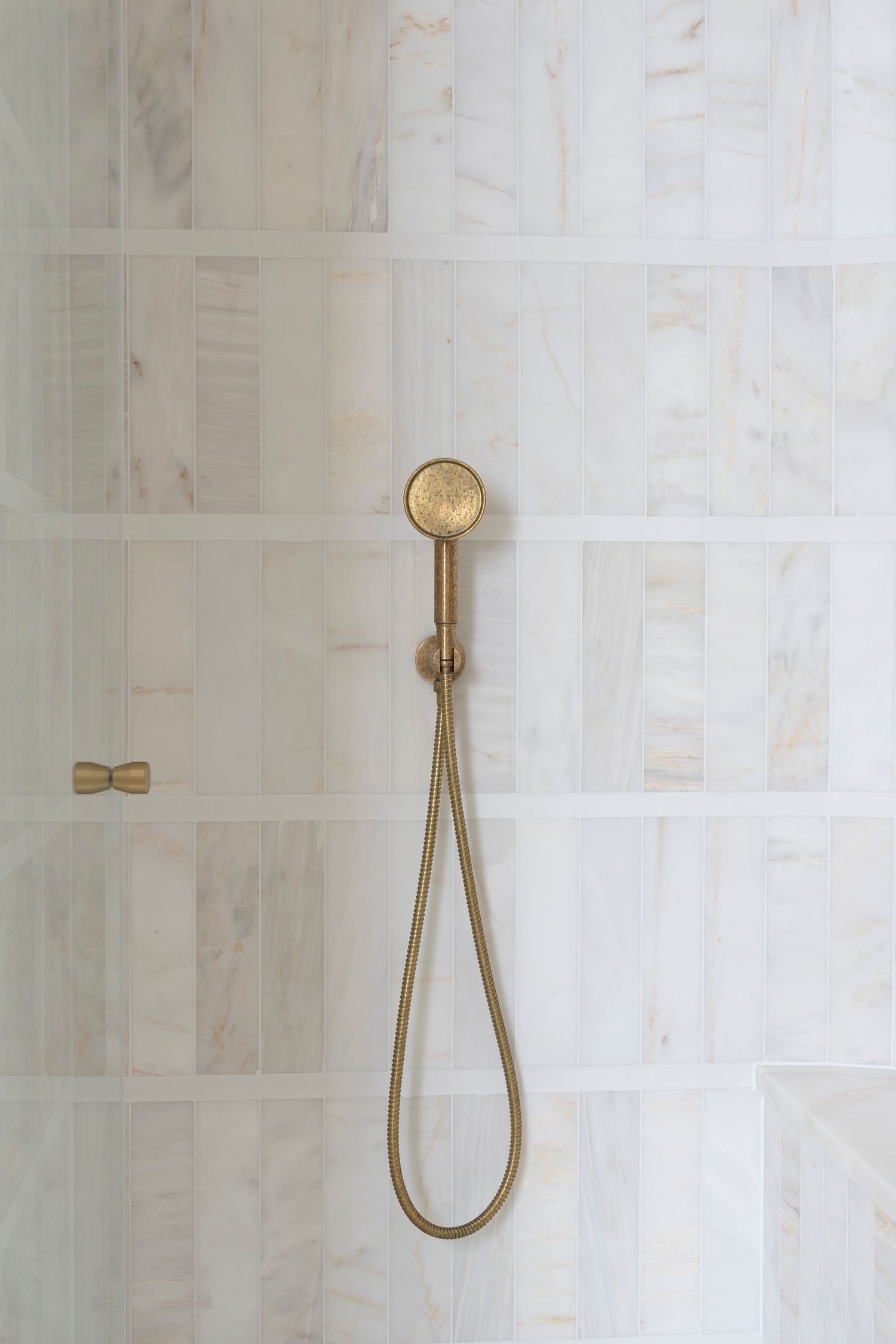 Close-up of brushed brass handheld shower head against light stone rectangular subway tiles.