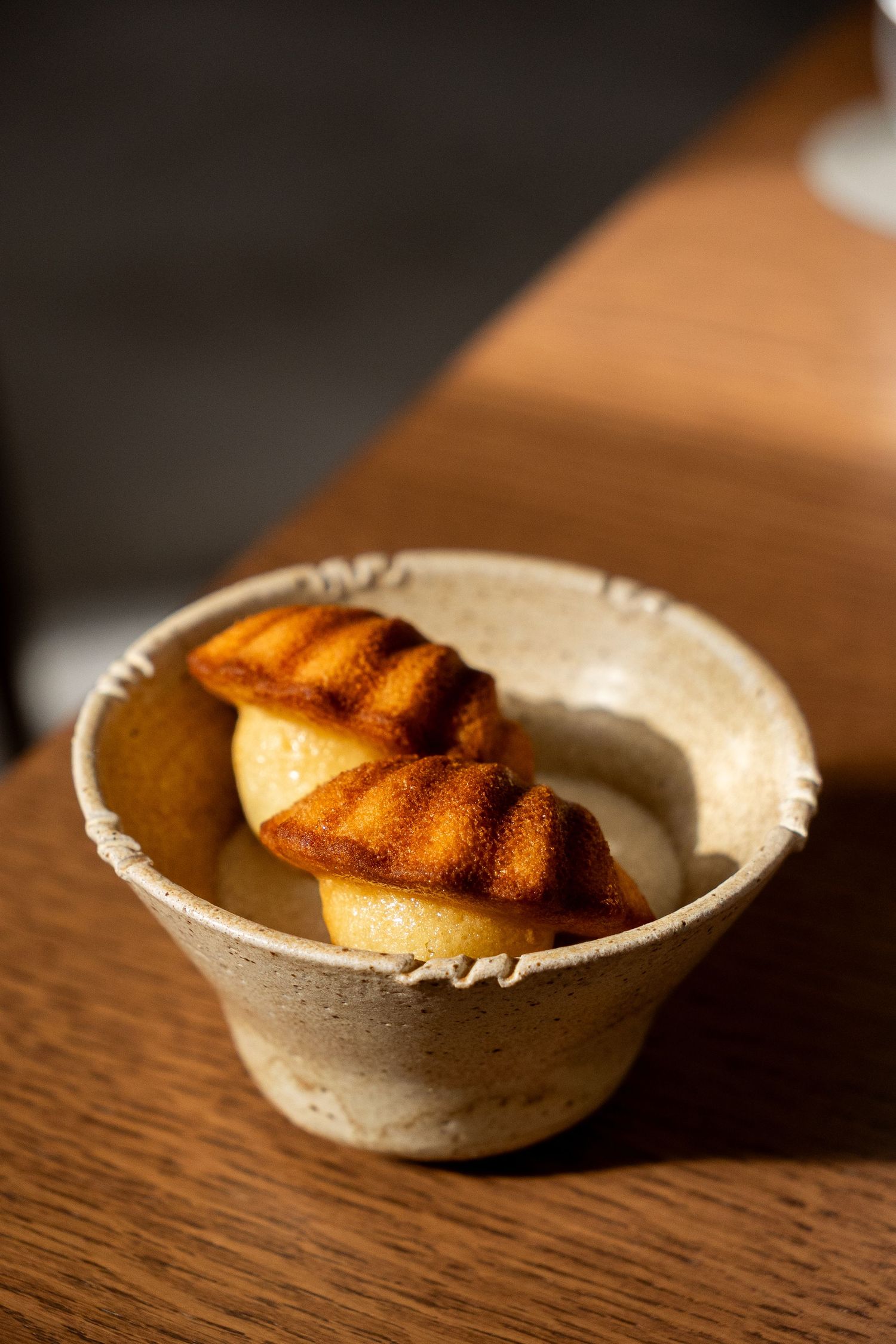 Golden brown madeleines served in a rustic ceramic bowl on a wooden table in warm sunlight.
