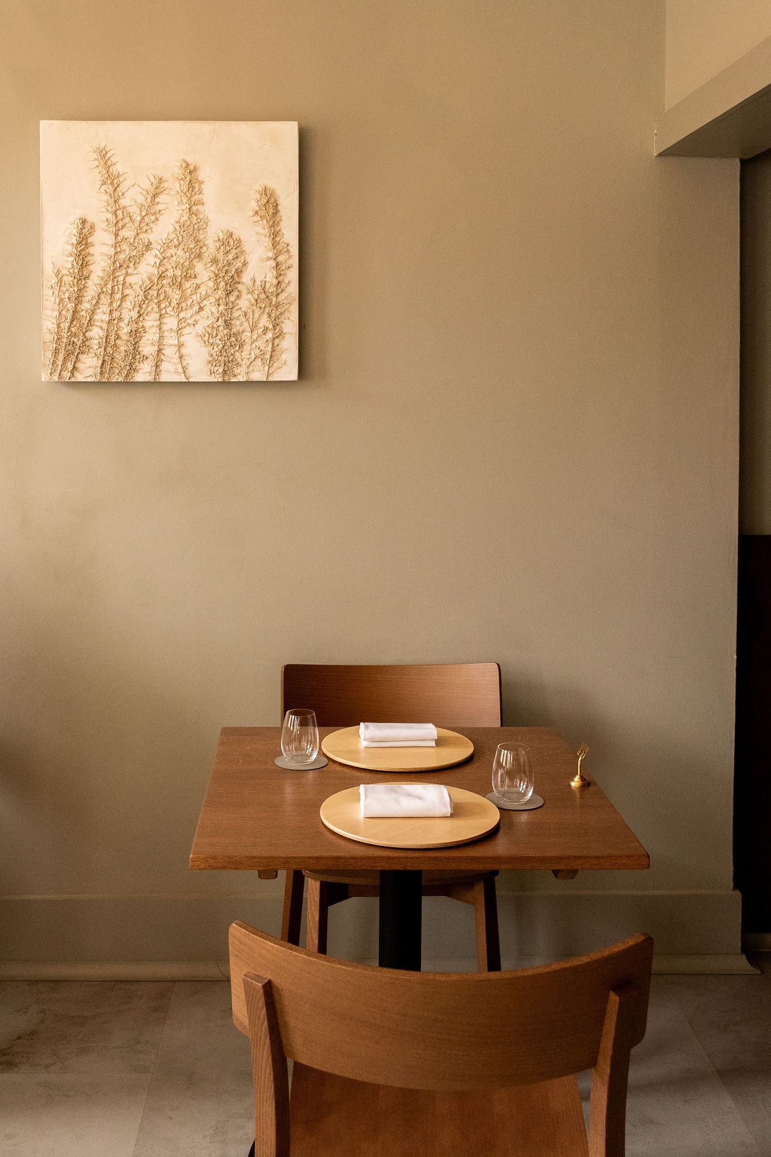Minimalist restaurant table set for two with wooden chairs, gold placemats, and botanical wall art.