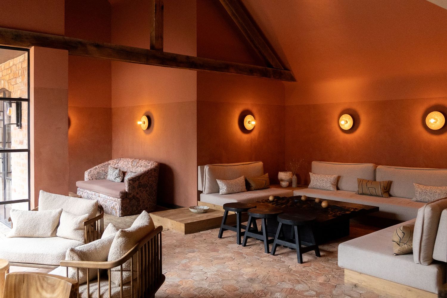 Rustic terracotta-walled lounge with vaulted ceiling, exposed beams, cream sofas, and circular gold wall sconces.
