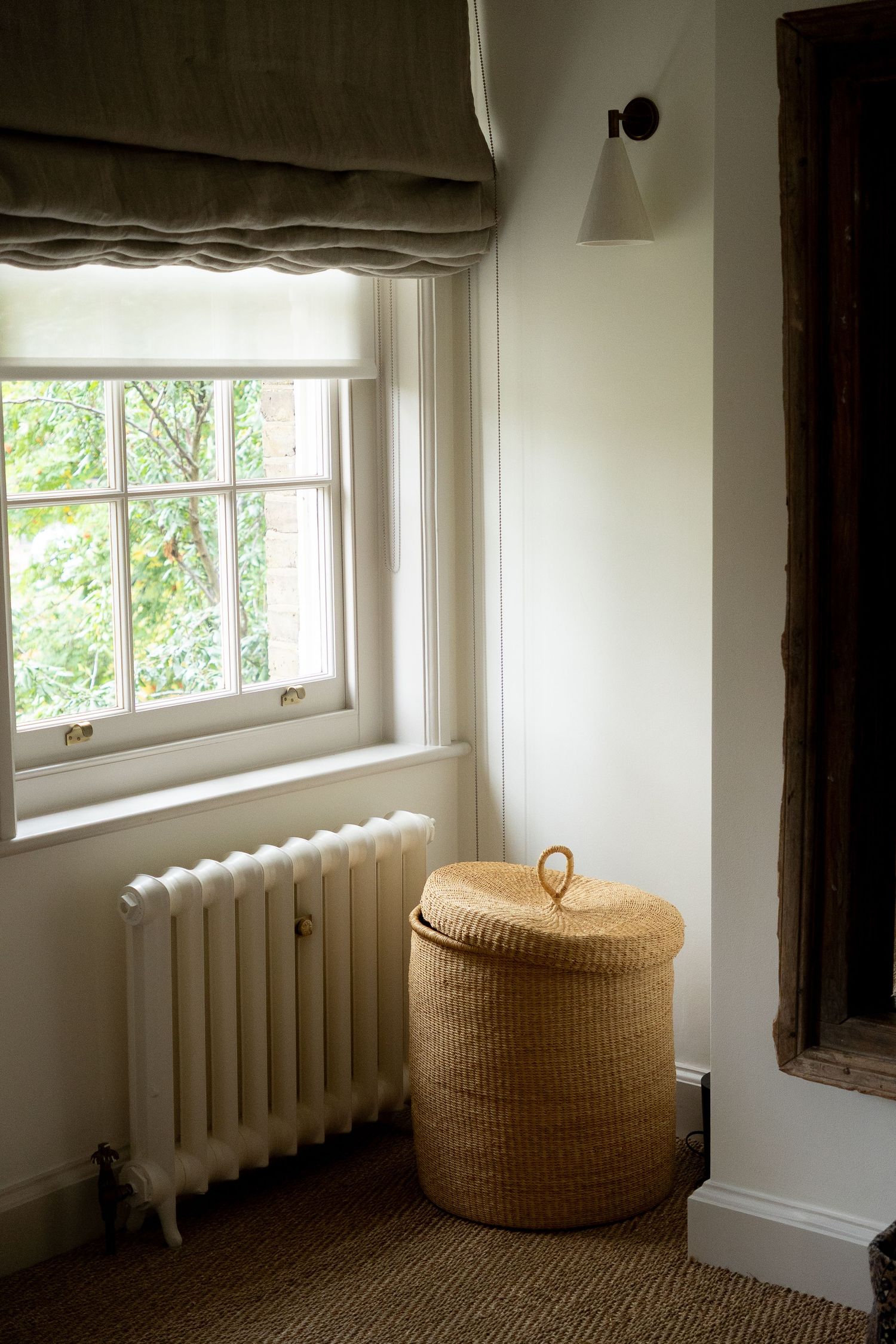 White cast iron radiator beside wicker basket under sash window with linen roman blind in cozy room.