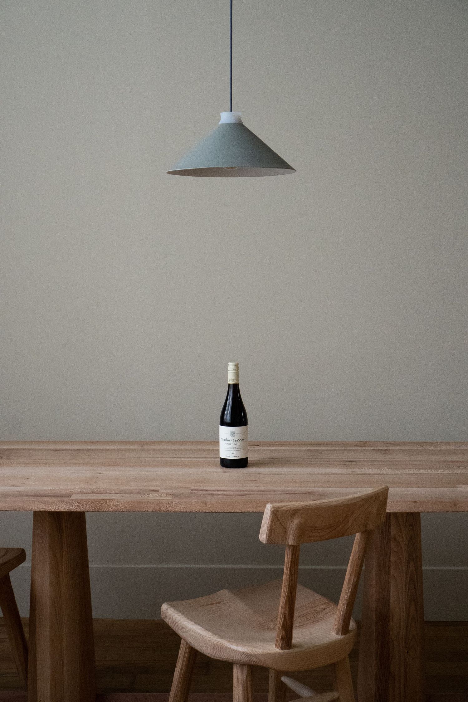 Minimalist dining room with wooden table, wine bottle, pendant light, and natural wood chair against sage green wall.