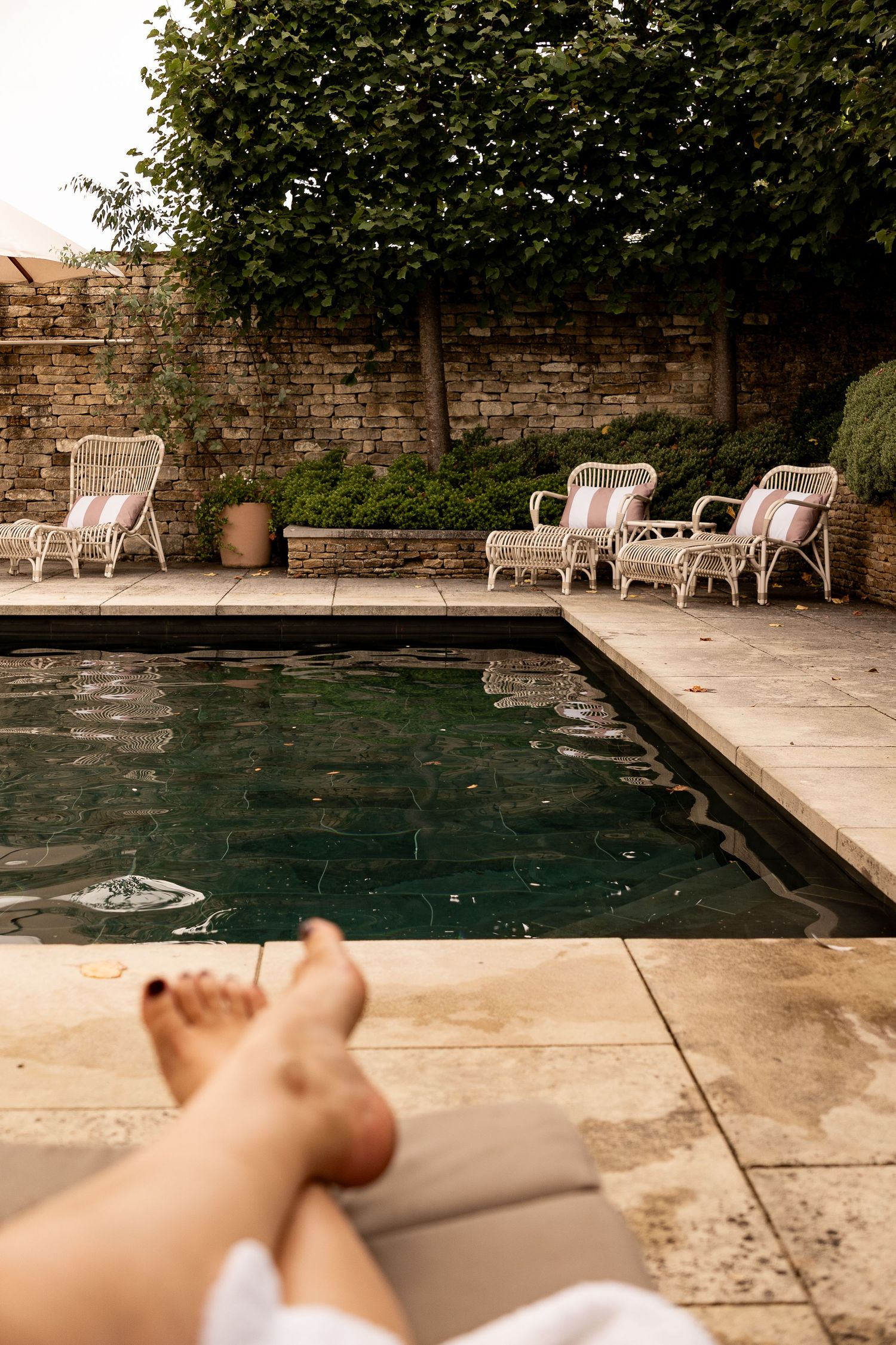 Relaxing poolside scene with person resting legs near dark water pool, rattan chairs, and stone garden wall.