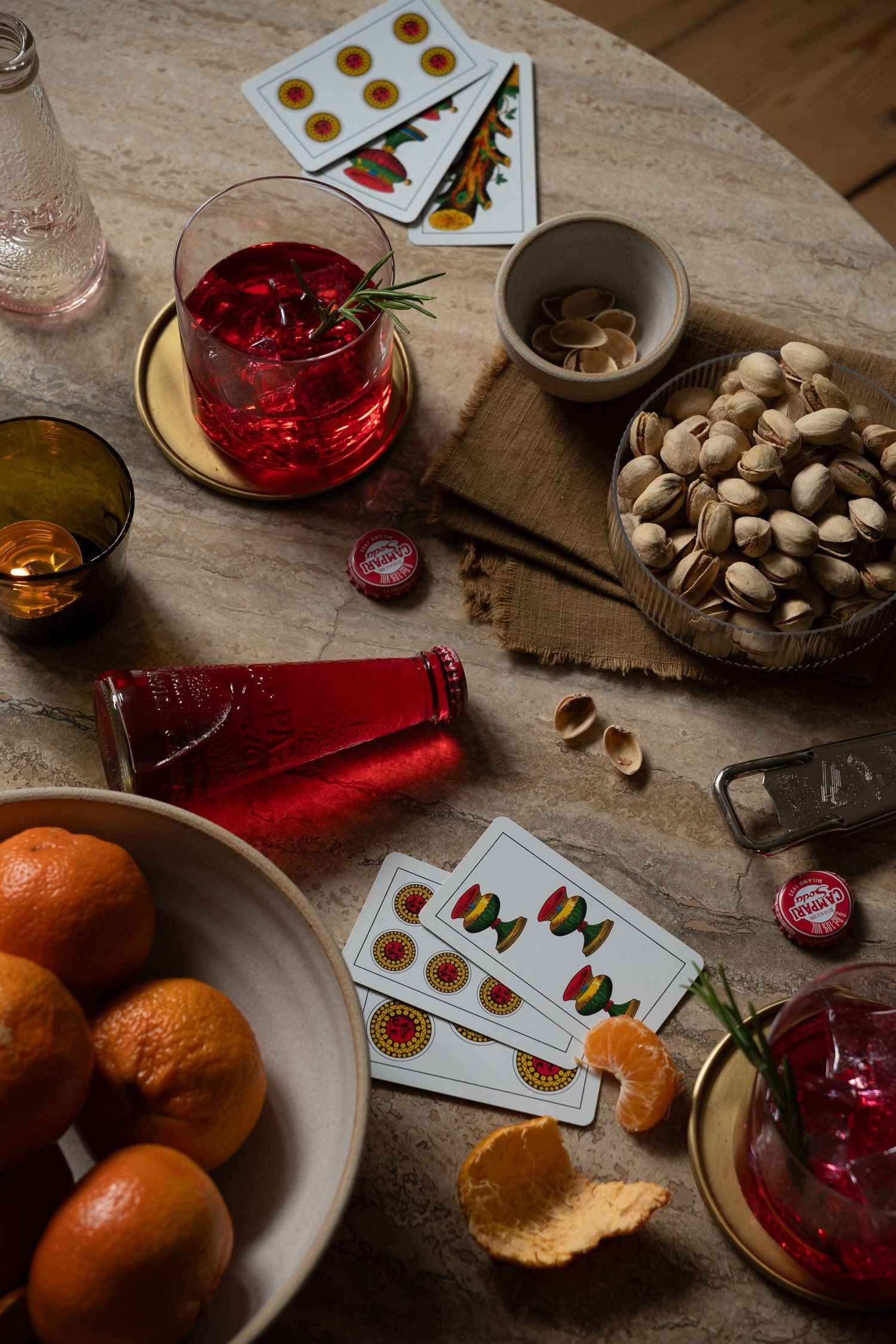 A marble table with playing cards, pistachios, oranges, and red cocktails for a cozy game night gathering.