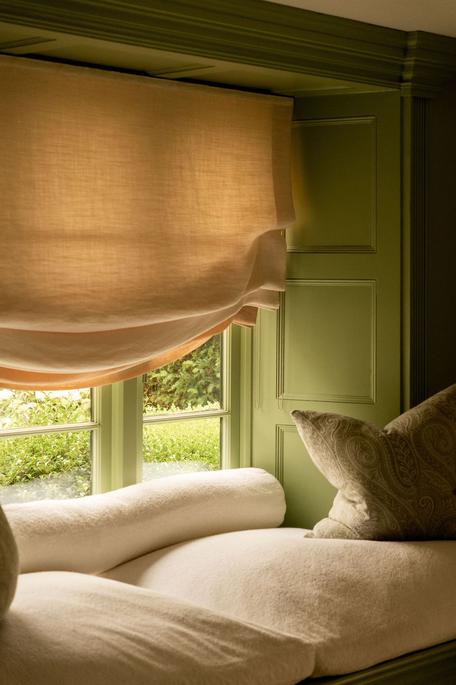 Cozy window seat with white cushions, linen roman blind, and green painted paneling overlooking garden greenery.